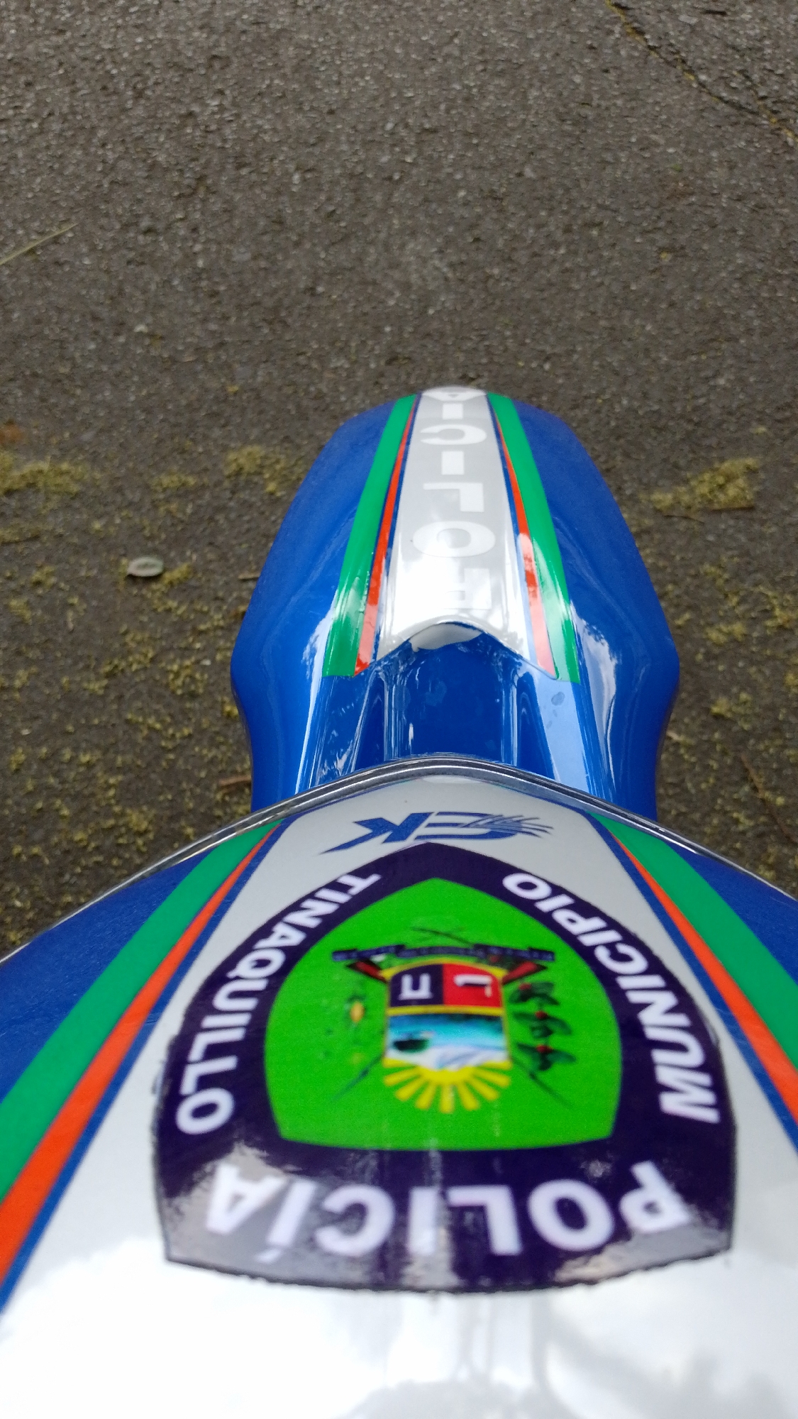 The image provides an overhead view of a blue and white motorcycle, likely a police vehicle, parked on a textured asphalt or concrete surface. In the immediate foreground, a part of the motorcycle, possibly a front fender or fairing, features a prominent circular decal. This decal clearly displays the white text "POLICIA MUNICIPIO TINAQUILLO" against a dark background, curved around a central emblem. The emblem itself depicts a landscape with a building, mountains, and a stylized sun. Flanking the decal and continuing onto the motorcycle's surface are vibrant green, white, and orange stripes.
Further back in the image, another section of the motorcycle, possibly the seat or rear fender, is visible. This part is also blue with a central white stripe bordered by green and orange stripes. The word "POLICIA" is printed vertically in white letters on the white stripe. The overall appearance suggests a police vehicle. The ground is grey and rough, indicative of a paved outdoor area. The glossy finish of the motorcycle parts reflects light, suggesting natural daylight. No specific activity is depicted beyond the stationary vehicle.