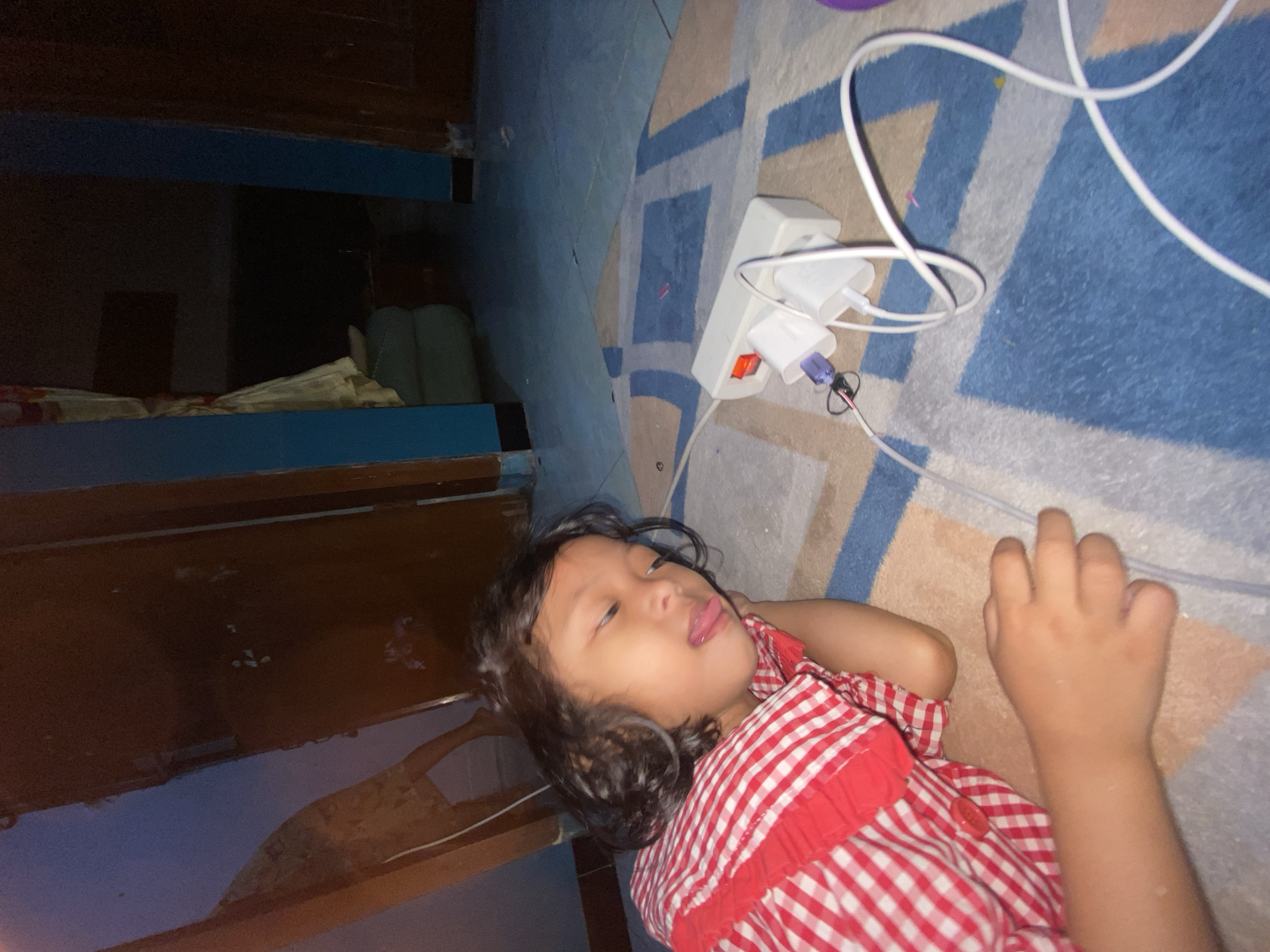 The image shows a young child lying on a carpeted floor, possibly in a bedroom. The child is wearing a red and white checkered shirt and has dark, curly hair. Their tongue is sticking out, suggesting playfulness or mischief. In the background, there's a power strip with several white adapters and cables, one of which is a white charging cable. The setting appears to be indoors, as indicated by the carpet and the presence of electrical outlets. The lighting is dim, suggesting it might be nighttime or a dimly lit room. The overall scene conveys a sense of a relaxed, everyday moment at home. There is no visible text in the image.