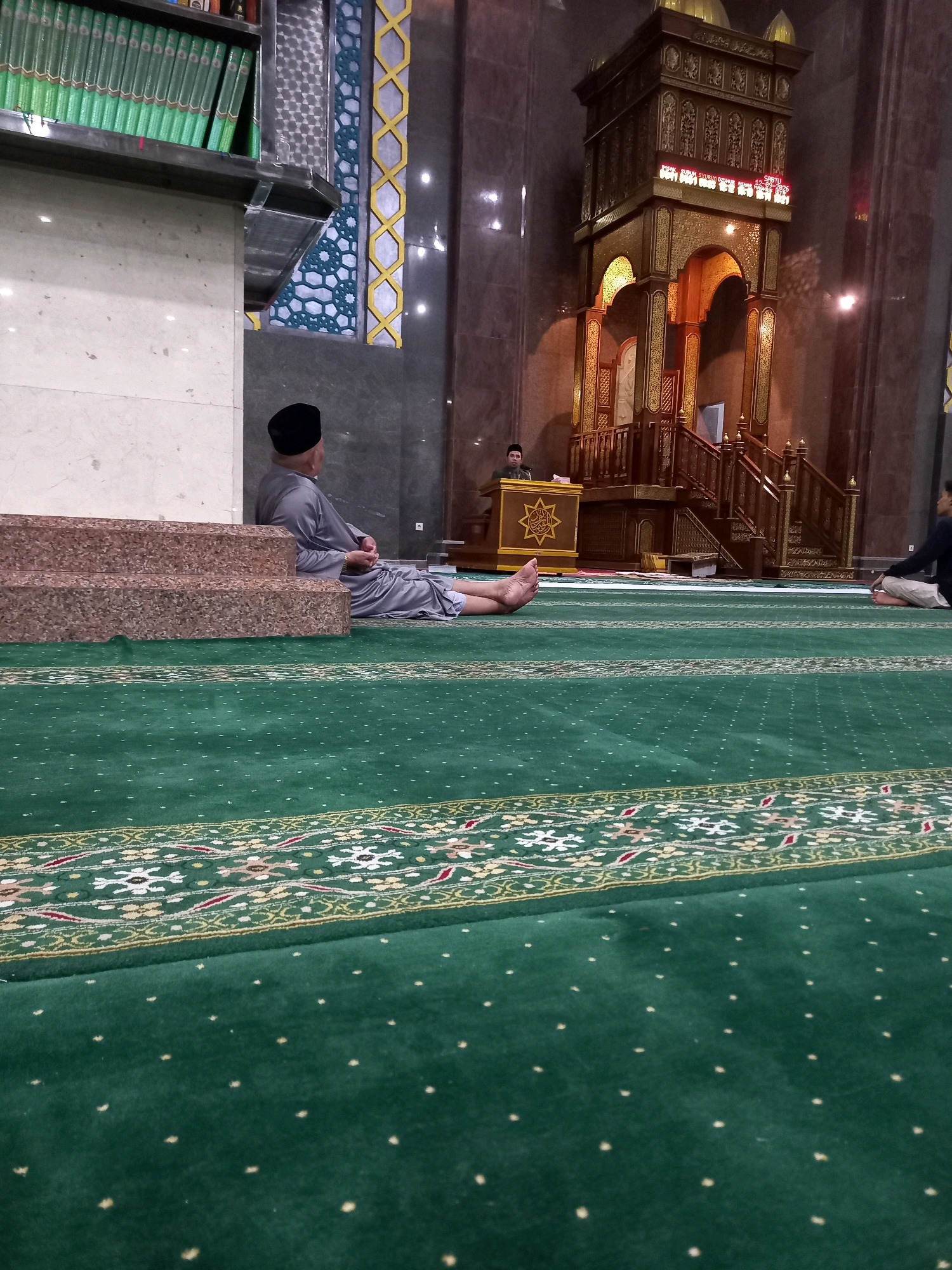 An interior shot taken inside a mosque shows a man in grey Islamic attire sitting on the floor with his legs extended, leaning against a stepped structure made of granite. He is wearing a black peci cap. The floor is covered with a green carpet featuring a patterned border. In the background, a wooden pulpit with ornate carvings and a digital display is visible, with another person standing behind it. The mosque's interior is illuminated by artificial lights, highlighting the brown marble walls and the intricate golden decorations of the pulpit. Bookshelves filled with green-bound books are positioned on the left side of the image, and decorative geometric patterns adorn the wall. The scene appears to be of a place of worship, likely in Samarinda, Indonesia, given the context provided.