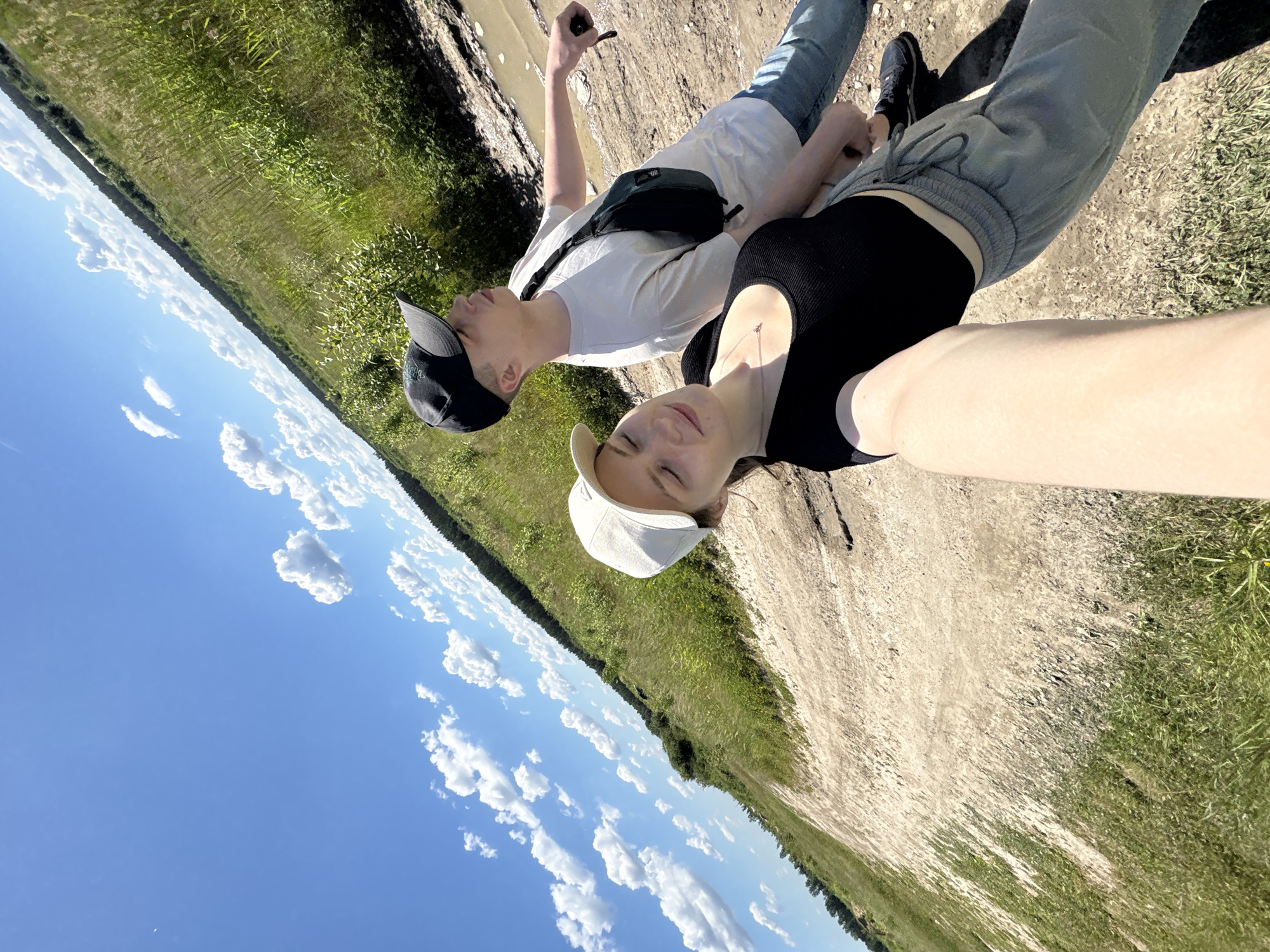 A young man and woman, appearing to be in their late teens or early twenties, are captured in a selfie-style photograph taken outdoors on a sunny day. The woman, positioned closer to the camera and slightly to the left, is wearing a white baseball cap, a black sleeveless top, and light grey sweatpants. She is looking towards the camera with her eyes closed and a faint smile. The man stands behind her and to the right, wearing a black baseball cap, a light grey t-shirt, and blue jeans. He has a dark fanny pack worn across his chest. His gaze is directed upwards and to the right.

They are standing on a dirt path with patches of dry grass and sparse vegetation. To their right, the path appears to transition into a muddy puddle. The background consists of a vast, grassy field with tall green reeds and scattered bushes, leading to a dense line of dark green trees in the distance. Above them, a bright blue sky is dotted with fluffy white cumulus clouds. The overall setting suggests a rural or natural environment, potentially on the outskirts of Manushkino, Russia, given the provided location information. The angle of the photograph and the presence of a muddy area could indicate they are on a trail or a less-maintained road. The lighting indicates it is daytime, likely late morning or early afternoon.