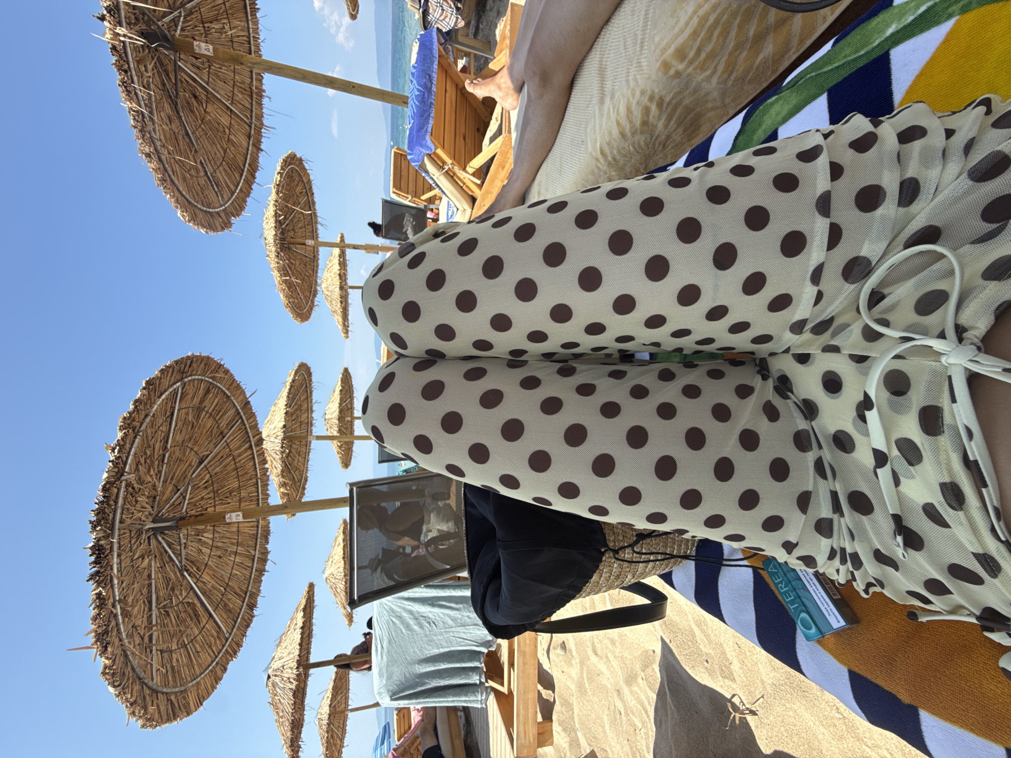On a sunny day at Ofrynio Beach, Greece, the perspective is from someone lying down, their legs covered in a sheer beige fabric with dark brown polka dots, positioned centrally in the foreground. The individual is on a beach towel that features stripes of blue, white, and orange. Next to their legs, a woven straw bag with a black interior and a black strap is visible, and beside it, a green and white pack of "TEREA" brand tobacco sticks lies on the towel.
Behind the person, the sandy beach stretches out, dotted with several straw umbrellas providing shade. To the left, a wooden beach lounger is covered with a light blue draped fabric. Further back, other beach loungers are visible, some occupied by unseen individuals. A framed picture displaying two people is propped up on one of the loungers. The background reveals the clear blue sky and the sparkling sea, indicating a typical beach scene. The general atmosphere is relaxed and summery.