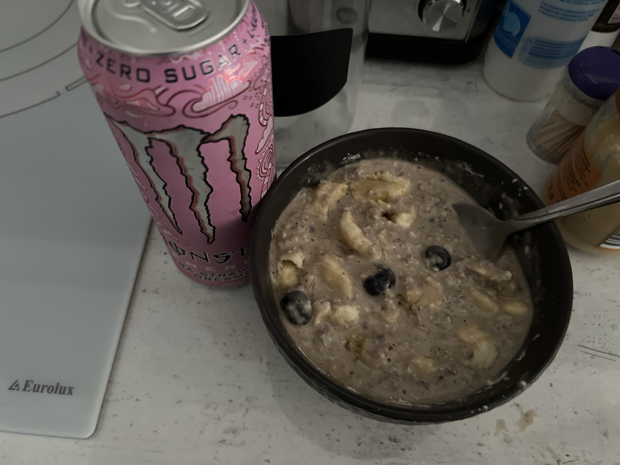 A close-up shot displays a can of pink Monster Zero Sugar energy drink next to a dark bowl filled with what appears to be oatmeal containing banana slices and blueberries. A spoon rests in the oatmeal. Background elements include a white cutting board, a black and silver appliance, and various containers. It is not possible to determine the location in the city of Burgas, Bulgaria, from this image.