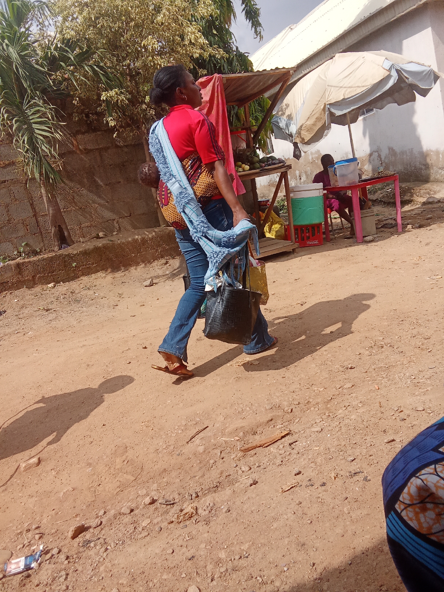 A woman, possibly of Nigerian descent, walks on a dirt road, carrying a baby on her back. She is wearing a red t-shirt, blue jeans, and sandals. A light blue and patterned cloth is draped over her shoulders and wraps around the baby. She also carries a black handbag in her right hand, and a yellow bag in her left hand. Her shadow is cast on the dusty ground, indicating it is a sunny day. In the background, a small market stall is visible, with an umbrella providing shade. Fruits and other goods are displayed on shelves. A person is seated at the stall, possibly a vendor or customer. A white building with a light-colored roof is behind the market. The scene appears to be in a rural or semi-urban area, consistent with Kagini, Nigeria. The overall mood is one of everyday life, with the woman going about her business.