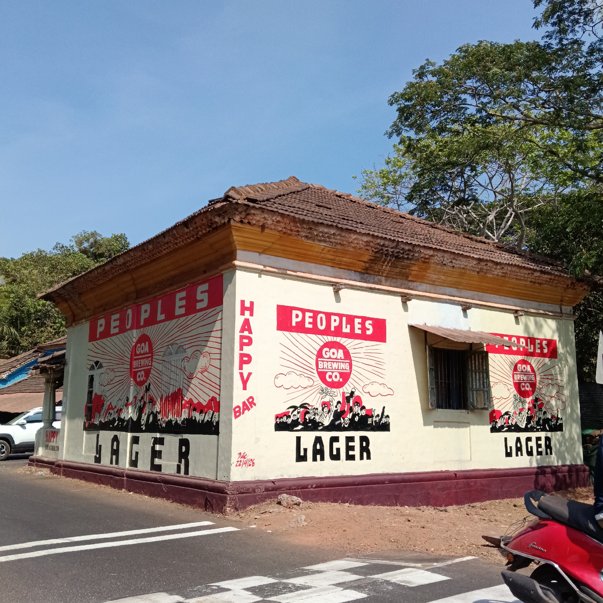 A daytime shot depicts a single-story building in Assagao, India, repurposed as a promotional canvas for "Peoples Brewing Co." The building's exterior walls are painted with the brewery's branding, featuring the words "PEOPLES," "GOA BREWING CO.," and "LAGER" in bold, contrasting colors. Red radial lines emanate from the brewery's logo, evoking a sense of energy. A "HAPPY BAR" sign adorns the side. A scooter parked on the road, and a nearby zebra crossing indicate the presence of vehicular and pedestrian traffic. The building's roof is tiled, and trees frame the scene, providing a backdrop of natural foliage. The sky is clear and blue, suggesting a sunny day.