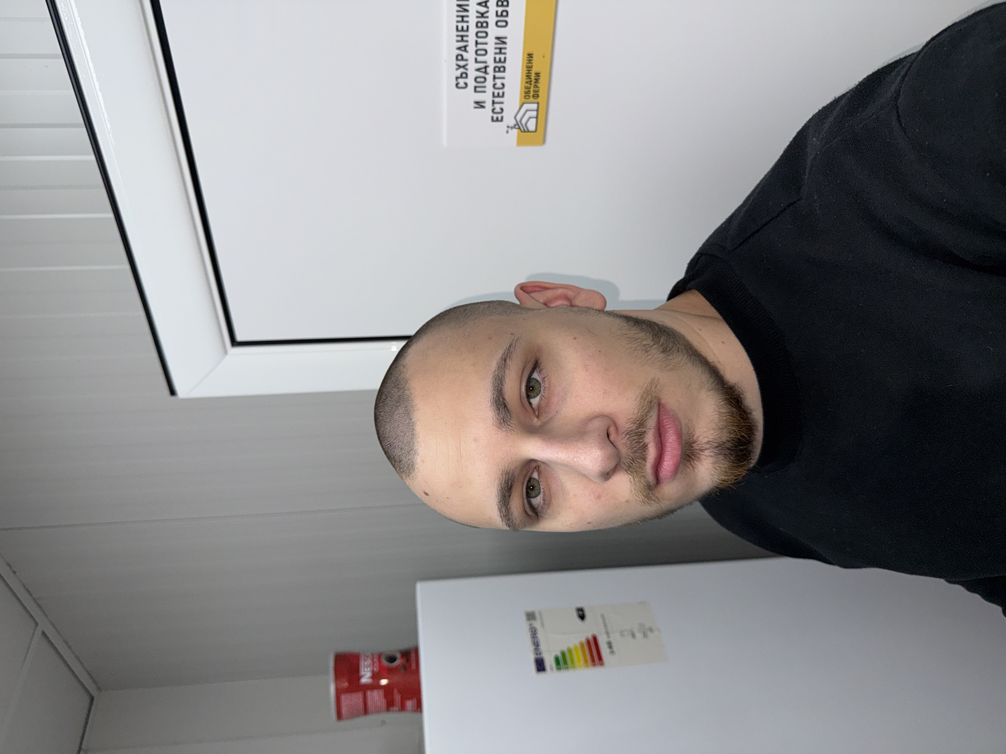 A medium shot depicts a man with light skin, short-cropped hair, and a trimmed beard. He's wearing a black long-sleeved shirt. The background features white walls, a white refrigerator with an energy efficiency sticker, and a red NESCAFE can. A sign in Bulgarian is visible on the upper wall; based on the sign and other visual elements, this image was taken in Stara Zagora, Bulgaria.