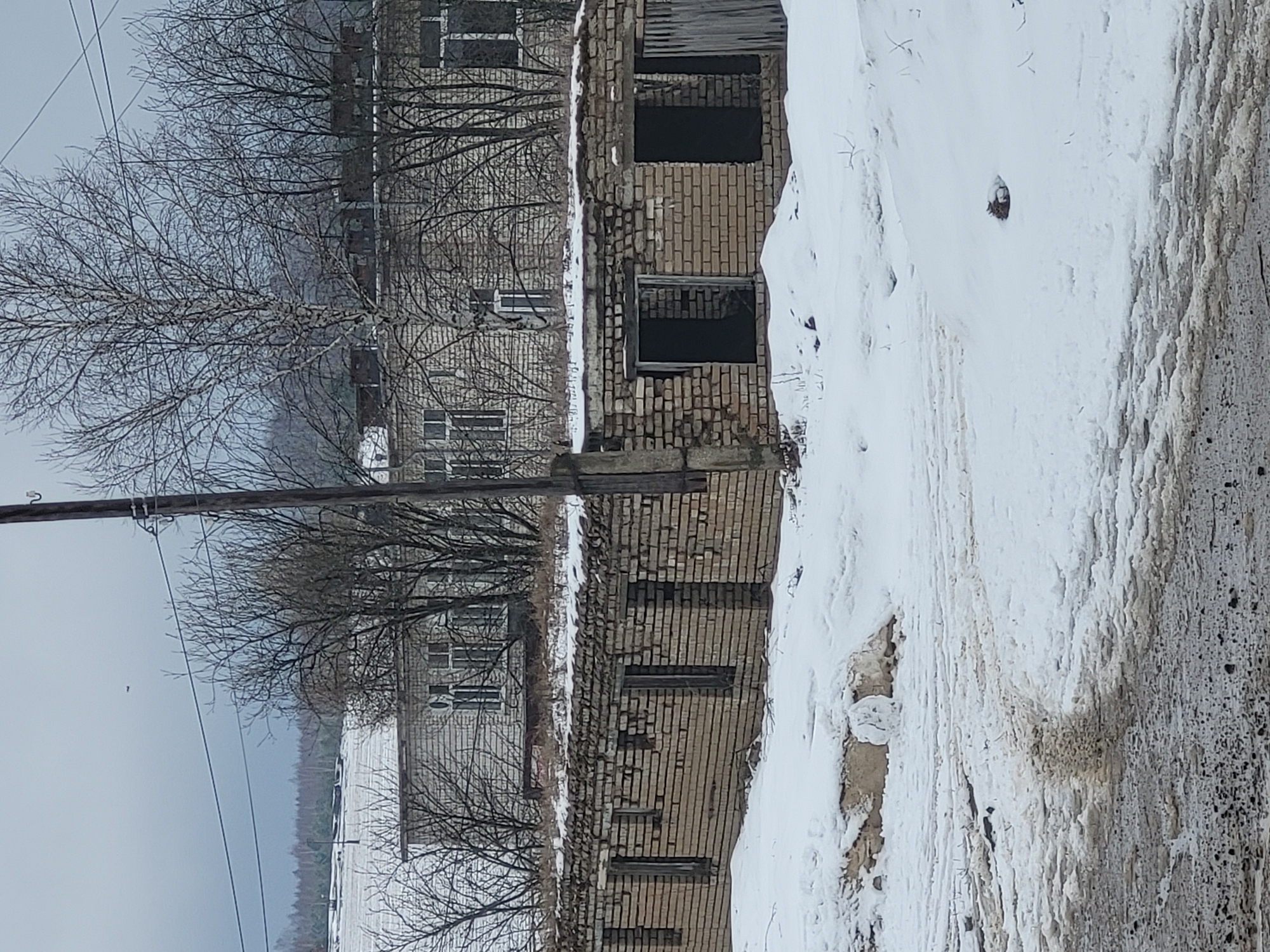 The image depicts a partially demolished brick building in a snowy environment, with the appearance of Зипуново, Russia. The building shows signs of decay and abandonment, with missing windows and crumbling brickwork. Snow covers the ground and parts of the building, indicating a cold, wintery setting. Bare trees are visible, reinforcing the season. An electrical pole and wires are in the foreground. In the background is a multi-storied building.