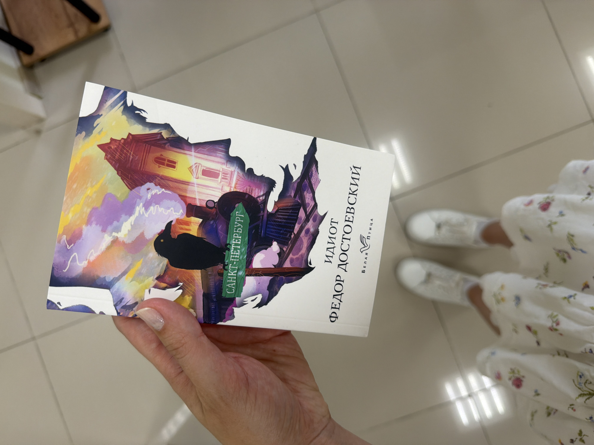 A woman holds a copy of Fyodor Dostoevsky's "The Idiot" book in her left hand. The book cover shows an illustration with colorful buildings, a silhouette of a man, and the title in Russian: "Идиот. Федор Достоевский." A green label reads "Санкт-Петербург" (Saint Petersburg). The woman wears a white dress with floral patterns. White sneakers are visible on her feet. The background consists of light gray tiles, suggesting an indoor location. The photograph does not contain any information that allows to determine the location in Rostov-on-Don.