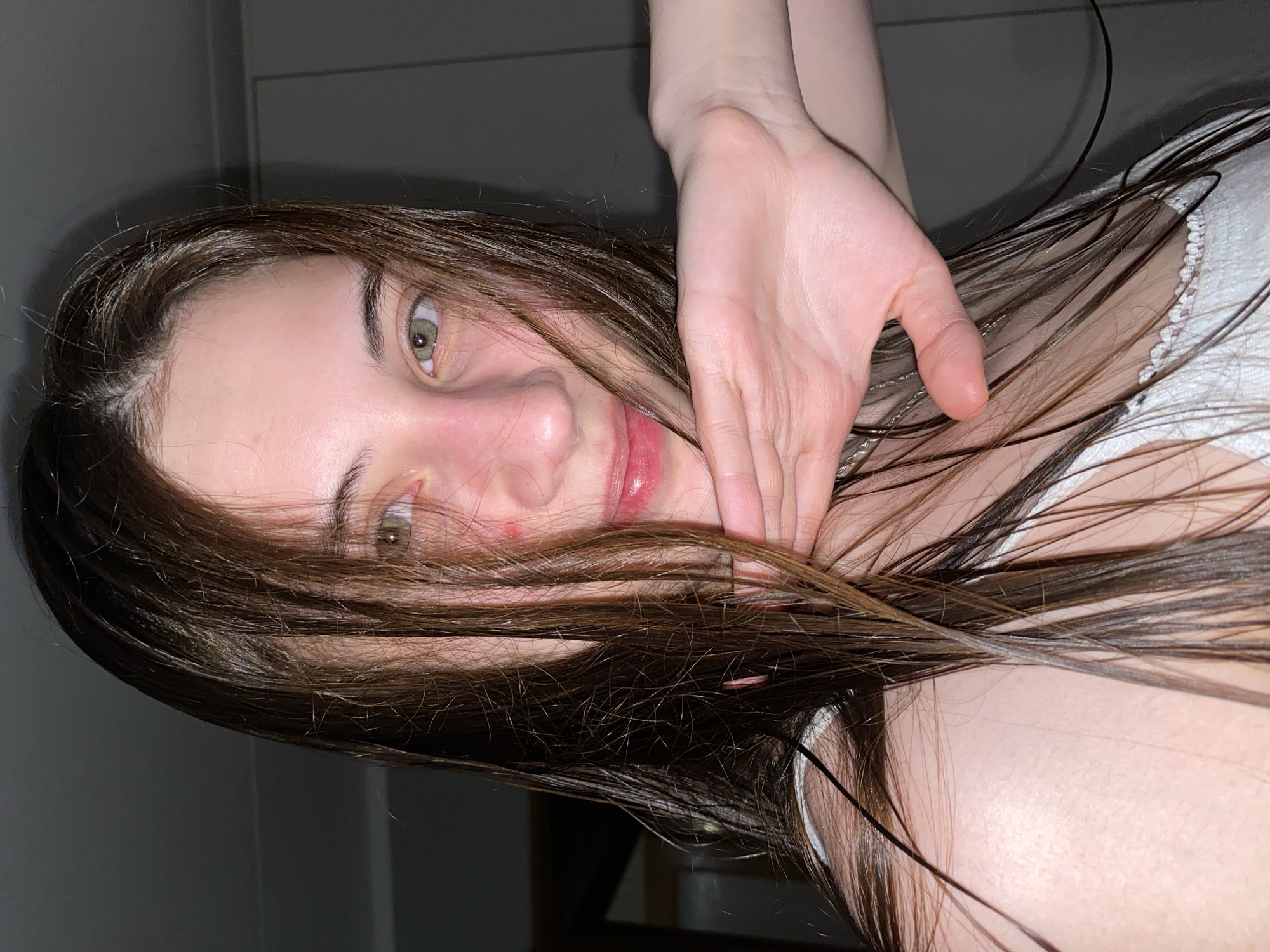 A young woman with light skin and hazel eyes, possibly in her late teens or early twenties, is looking at the camera with a slight smile. Her long, wet, dark brown hair is partially covering her face and shoulder. She has a red mark under her nose and red lipstick on her lips. A hand is positioned near her neck. She appears to be wearing a white top with lace trim. The background is a gray wall, suggesting an indoor setting. There is no visual information in this image to suggest the location is Votkinsk, Russia.