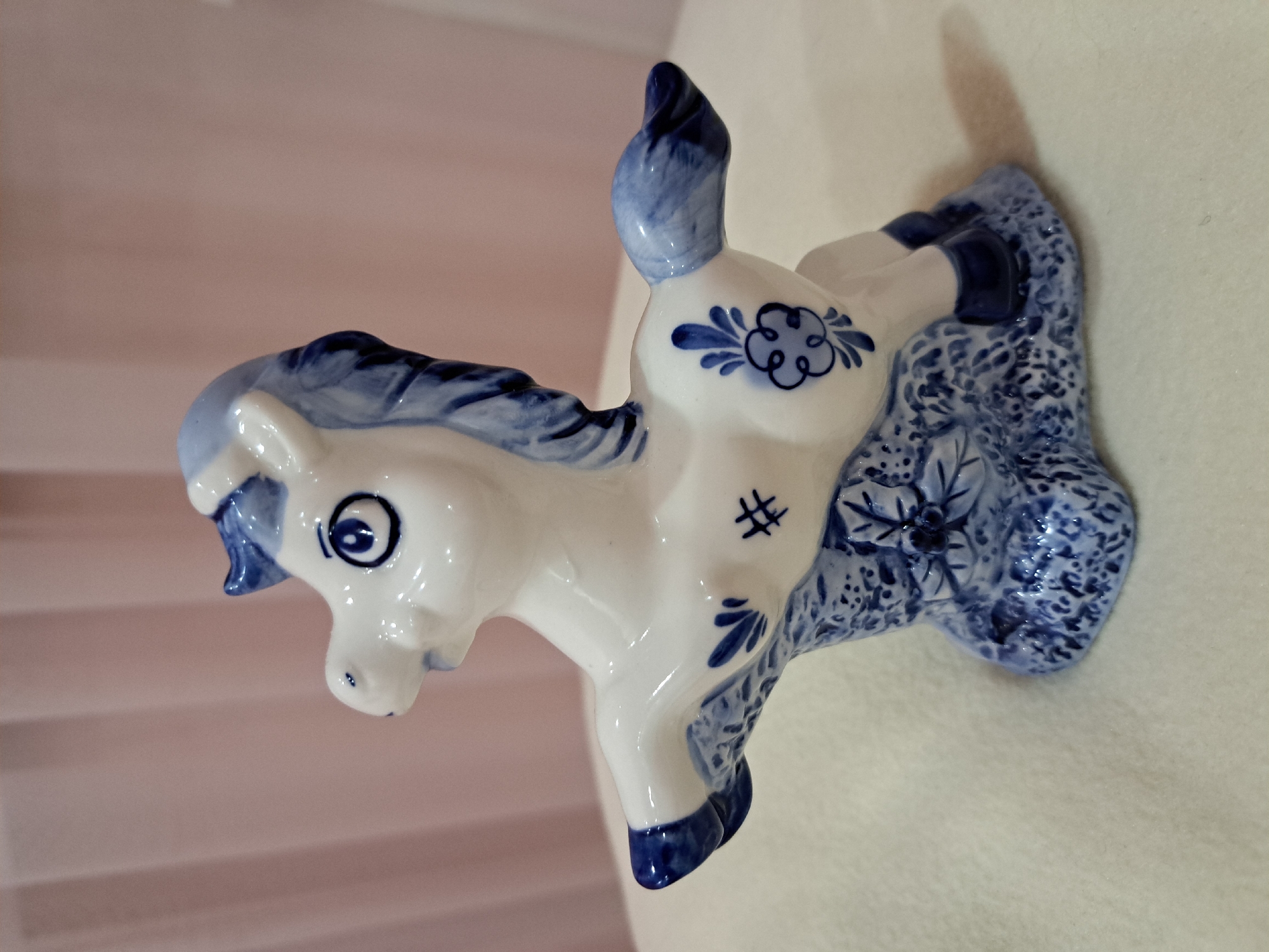 A white and blue porcelain figurine of a horse is displayed on a light-colored surface against a pink, draped fabric background. The figurine is decorated with intricate blue floral patterns and has a stylized mane and tail. The horse's front legs are extended as if in mid-stride, and it stands on a textured base adorned with similar blue designs. The overall impression is one of delicate craftsmanship and traditional artistry. The provided image does not contain information regarding its location within Irkutsk, Russia.