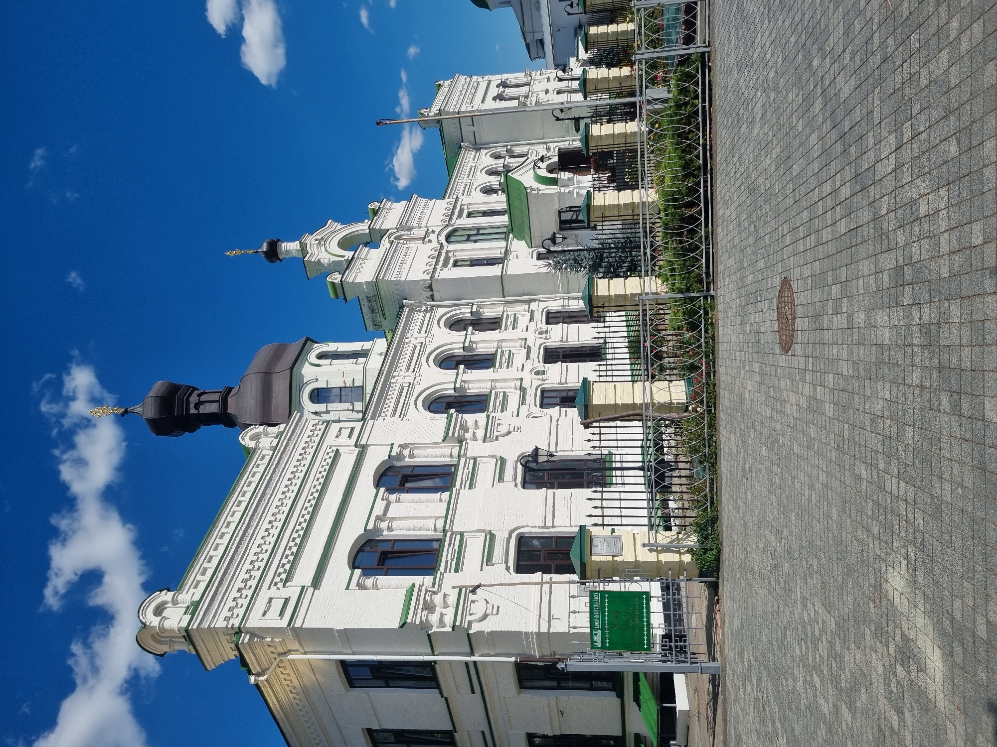 The image shows the exterior of a white brick building with green trim and roofs, likely a church or monastery, under a bright blue sky with scattered white clouds. The building features ornate architectural details, including arched windows, decorative moldings, and domes topped with crosses. A metal fence with decorative elements encloses the front of the property, separating it from a paved courtyard. A green sign with text in Cyrillic is visible near the entrance gate. The building's facade suggests historical significance. Based on its architectural style and the presence of Cyrillic script, this building is likely located in Kyiv, Ukraine, possibly a religious or historical site.