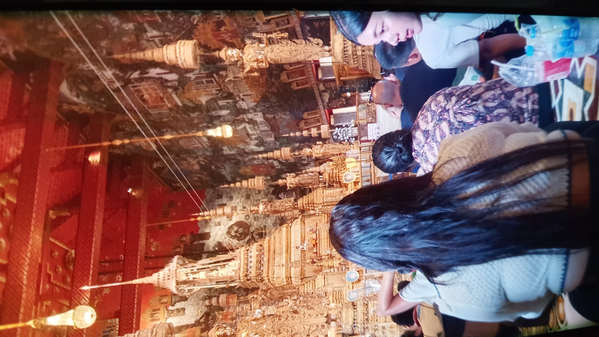 This media file depicts an indoor scene, likely a temple or religious site, given the ornate golden structures resembling stupas and intricate murals adorning the walls and ceiling. The image is oriented sideways, with the people appearing to be standing or sitting, looking upwards at the detailed artwork.
Several individuals are visible in the foreground and mid-ground. One person in the top right is wearing a white top and looking towards the ceiling, with a water bottle and plastic bag nearby. Another individual, dressed in a patterned garment, is seated further down. The closest subject, on the right, has long dark hair and is wearing a light-colored knitted top. A few other individuals are partially visible, suggesting a small group observing the surroundings.
The overall atmosphere appears calm, with the focus of the people directed towards the elaborate decorations. The lighting suggests it could be daytime, but the interior setting makes it difficult to determine definitively. There are no clear indicators of weather.
The location is suggested to be Koh Phangan, Thailand, which aligns with the architectural style and religious imagery often found in Thai temples. No visible text is discernible in the image. The prominent golden structures and detailed murals are the most notable details of the scene, creating a visually rich and culturally significant environment.