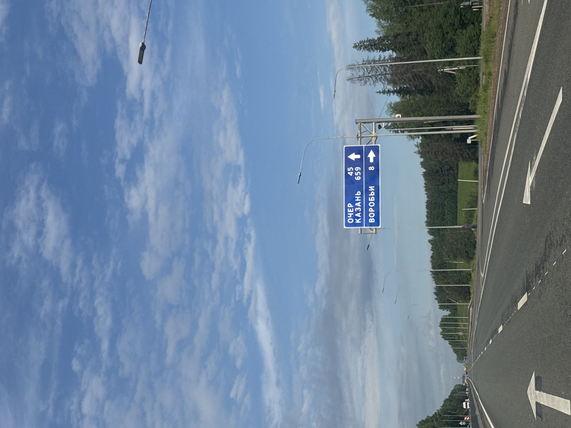 This landscape-oriented image captures a bright daytime scene on a road near Nytva, Russia, under a clear blue sky with scattered white clouds. The asphalt road, marked with white lines and a large white arrow indicating a turn or exit to the right, stretches into the distance, lined by numerous streetlights. On the right side of the road, green trees and foliage are visible, suggesting a rural or suburban highway setting.
The primary subject is a blue highway sign with white text and arrows, indicating directions and distances to various destinations. The sign is mounted on a metal gantry above the road. The visible text in Russian reads:
* An arrow pointing to the left next to "ОЧЕР" (OCHER) and the number "45".
* Below that, another arrow pointing to the left next to "КАЗАНЬ" (KAZAN) and the number "659".
* At the bottom, an arrow pointing to the right next to "ВОРОБЬИ" (VOROBYI) and the number "8".
These numbers likely represent distances in kilometers to the respective towns or cities. The bright lighting suggests it is mid-day, and the weather appears clear and sunny. No people or specific activity are distinctly visible, but the scene implies travel along a highway.