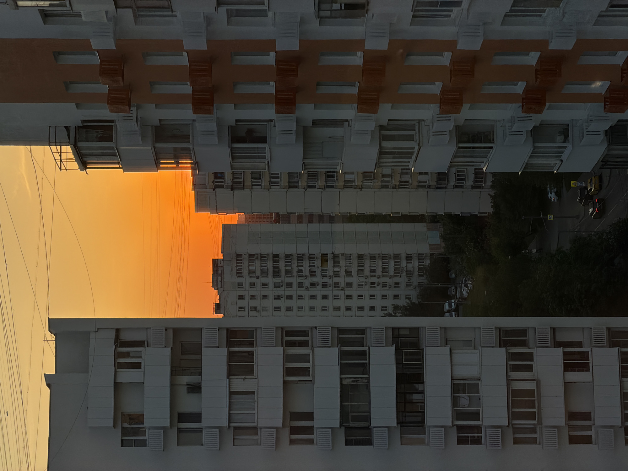 The image depicts a view between two high-rise buildings in Khimki, Russia. The buildings, predominantly white with some orange accenting on the upper one, frame an orange-hued sky, suggestive of either sunset or sunrise. Another, taller building is visible further in the distance. Below, there is a street with cars and trees.