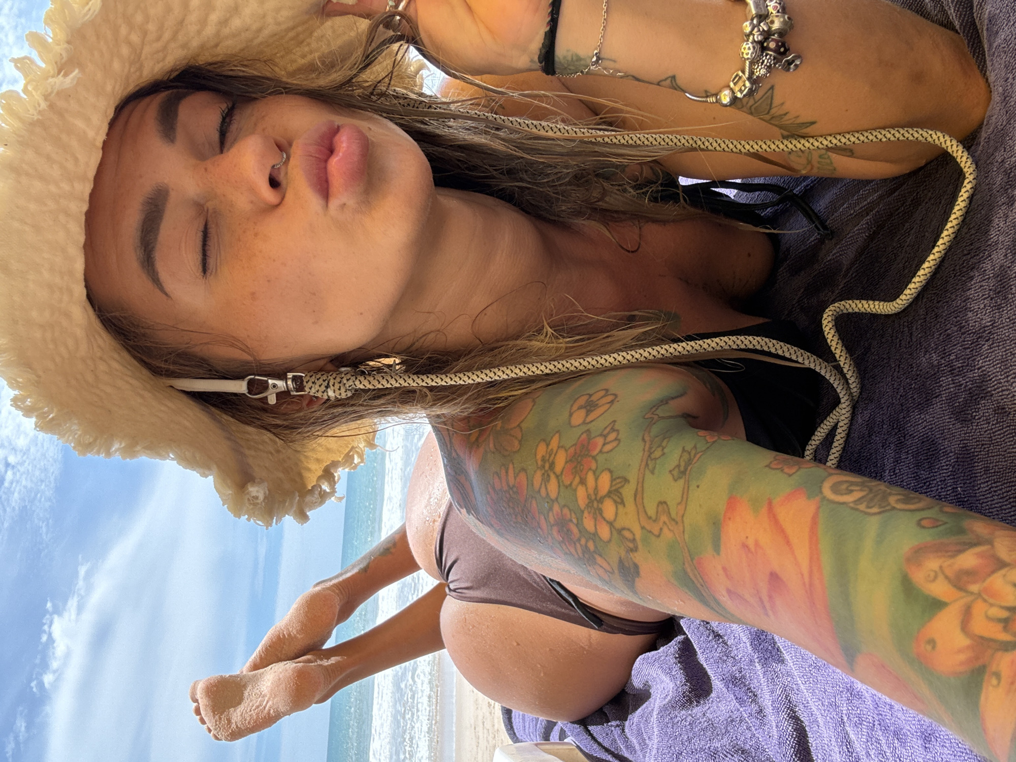 A woman with a large floral tattoo on her arm and leg is lying on a purple towel on a beach. She is wearing a brown bikini and a straw hat. Her eyes are closed and she is puckering her lips. Her feet are in the air, with sand on them. The ocean and a blue sky with clouds are visible in the background. The image appears to have been taken in Choeng Thale, Thailand.