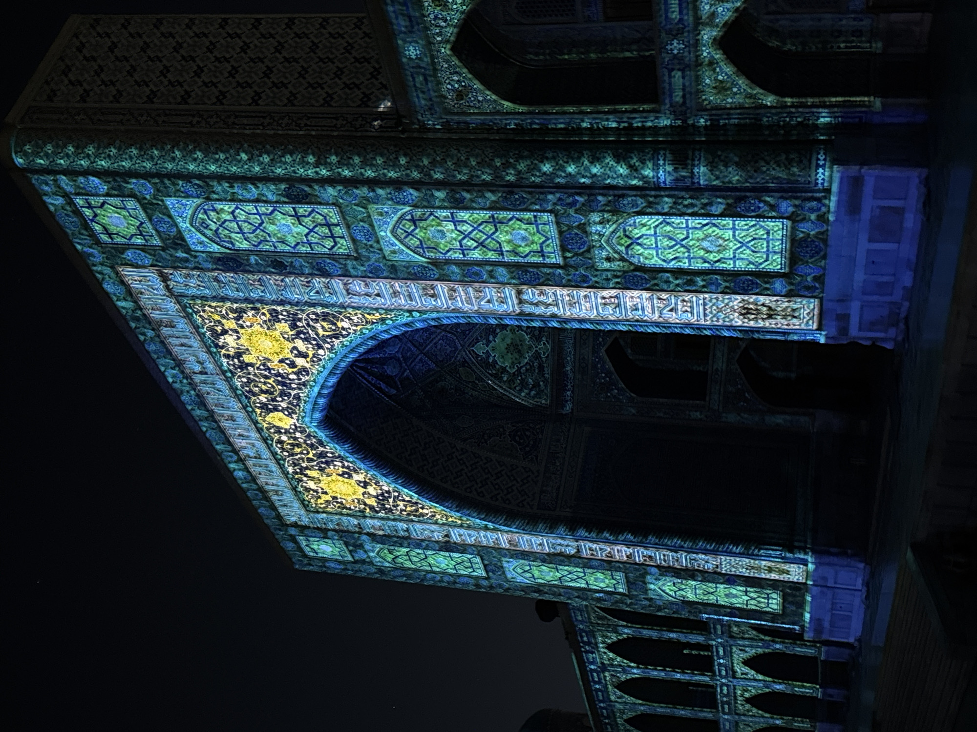 The image shows a low-angle view of a portion of a building at night, likely a facade or entrance, possibly in Samarkand City, Uzbekistan, judging by the architecture. The visible structure is intricately decorated with mosaic tiles in shades of blue, green, and yellow. There's an arched opening into a darker interior space. The decorative elements include geometric patterns, floral motifs, and possibly some inscriptions. Light illuminates the facade, highlighting the colors and textures of the tiles. The surrounding environment appears dark and undefined.