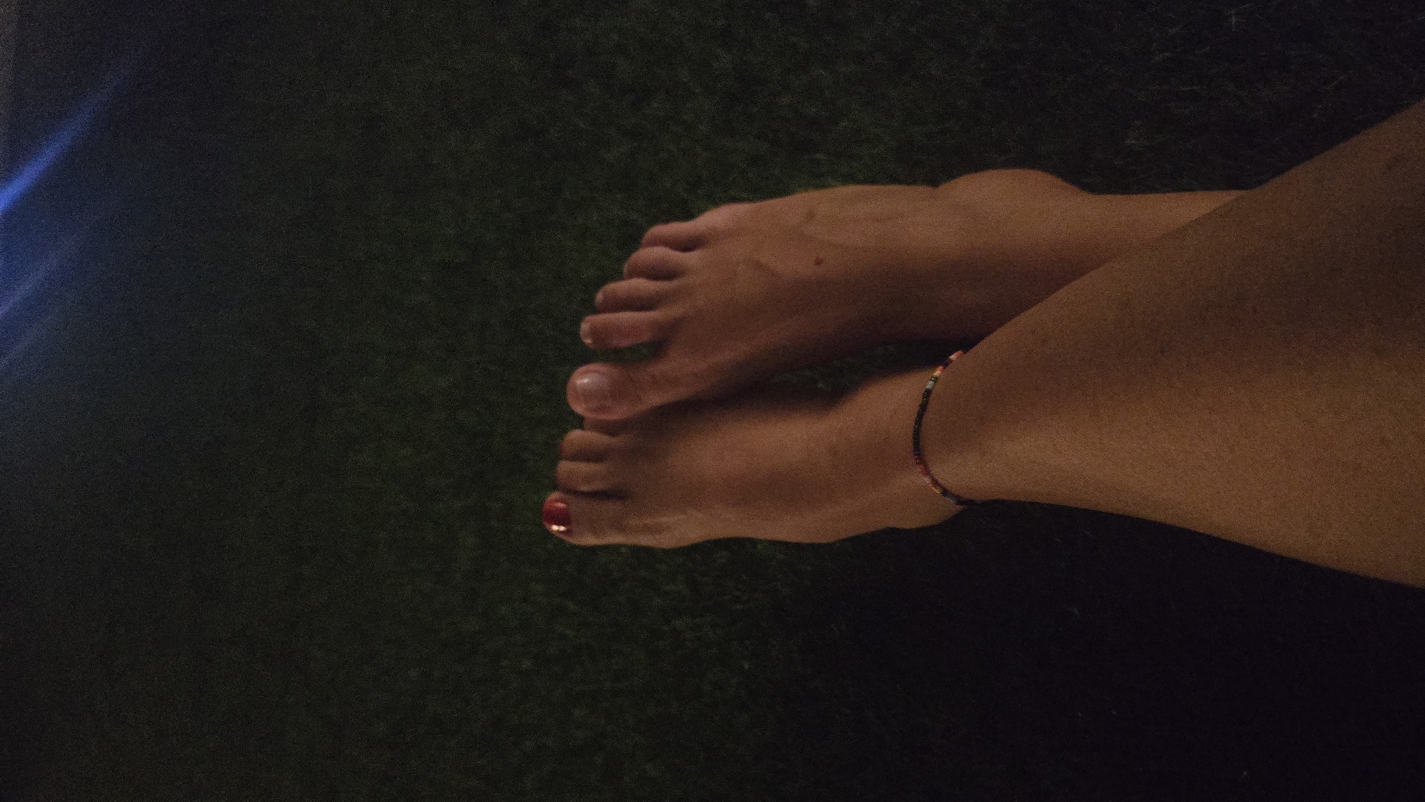A close-up, low-angle shot shows two bare feet resting on a dark, grassy surface at night. The feet are crossed, with the left foot positioned over the right. The toenails of the left foot are painted a vibrant red. A beaded anklet, featuring a mix of colored beads, adorns the left ankle. The background is dark and out of focus, with some bluish light streaks visible in the upper right corner, suggesting artificial lighting. The overall impression is one of relaxation and casual repose in an outdoor setting.