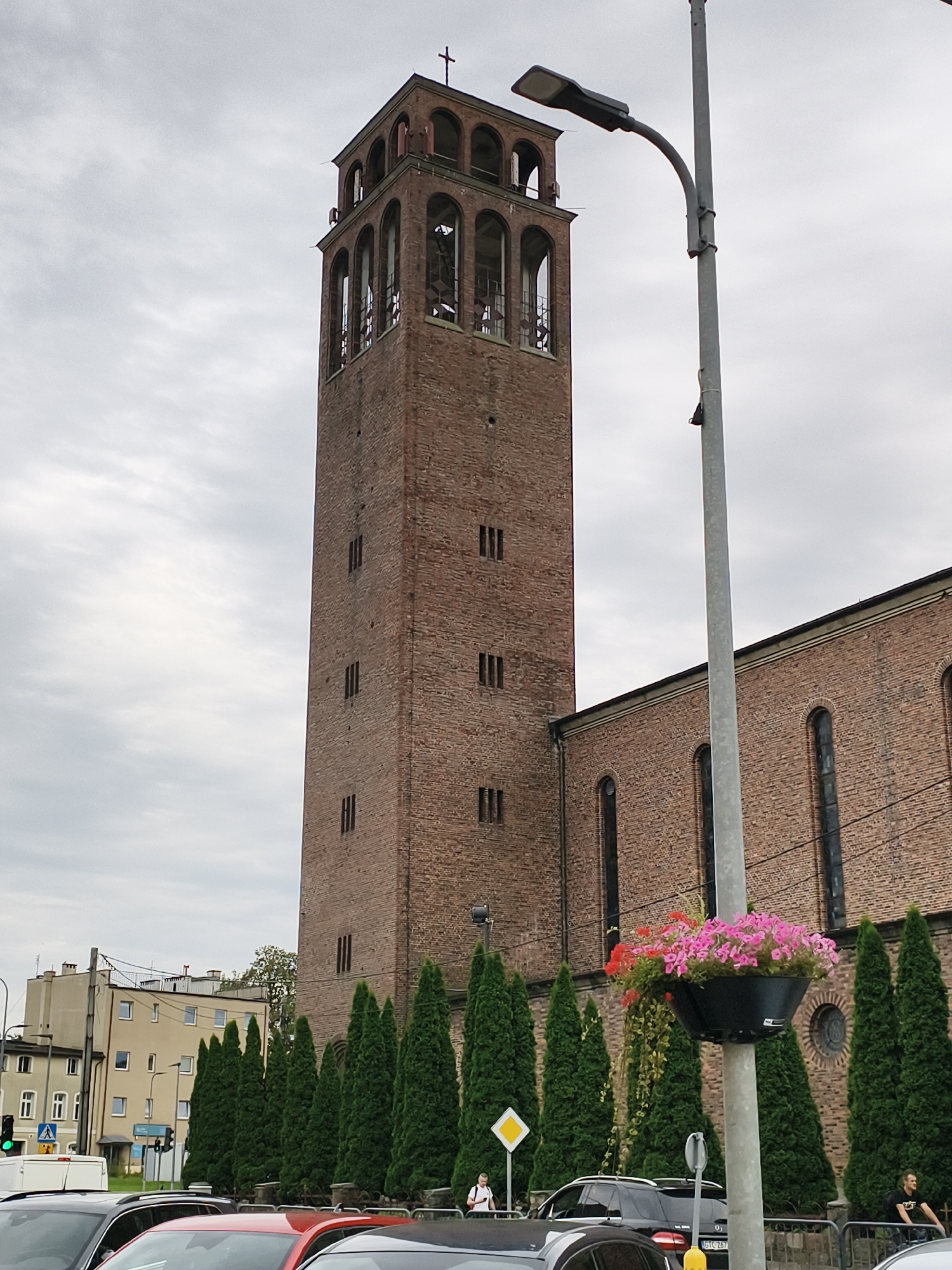 The image shows a view of the Holy Heart of Jesus Church in Tczew, Poland, on a cloudy day. The church features a tall, red brick bell tower with arched openings at the top and a cross on its peak. Adjacent to the tower is the main body of the church, also constructed of red brick with arched windows. In the foreground, a street lamp with a flower pot filled with pink and red flowers is visible. Several evergreen trees are planted in a row in front of the church. The street in front of the church is lined with cars, and a few people are seen standing or walking on the sidewalk. Buildings and traffic lights are visible in the background to the left.