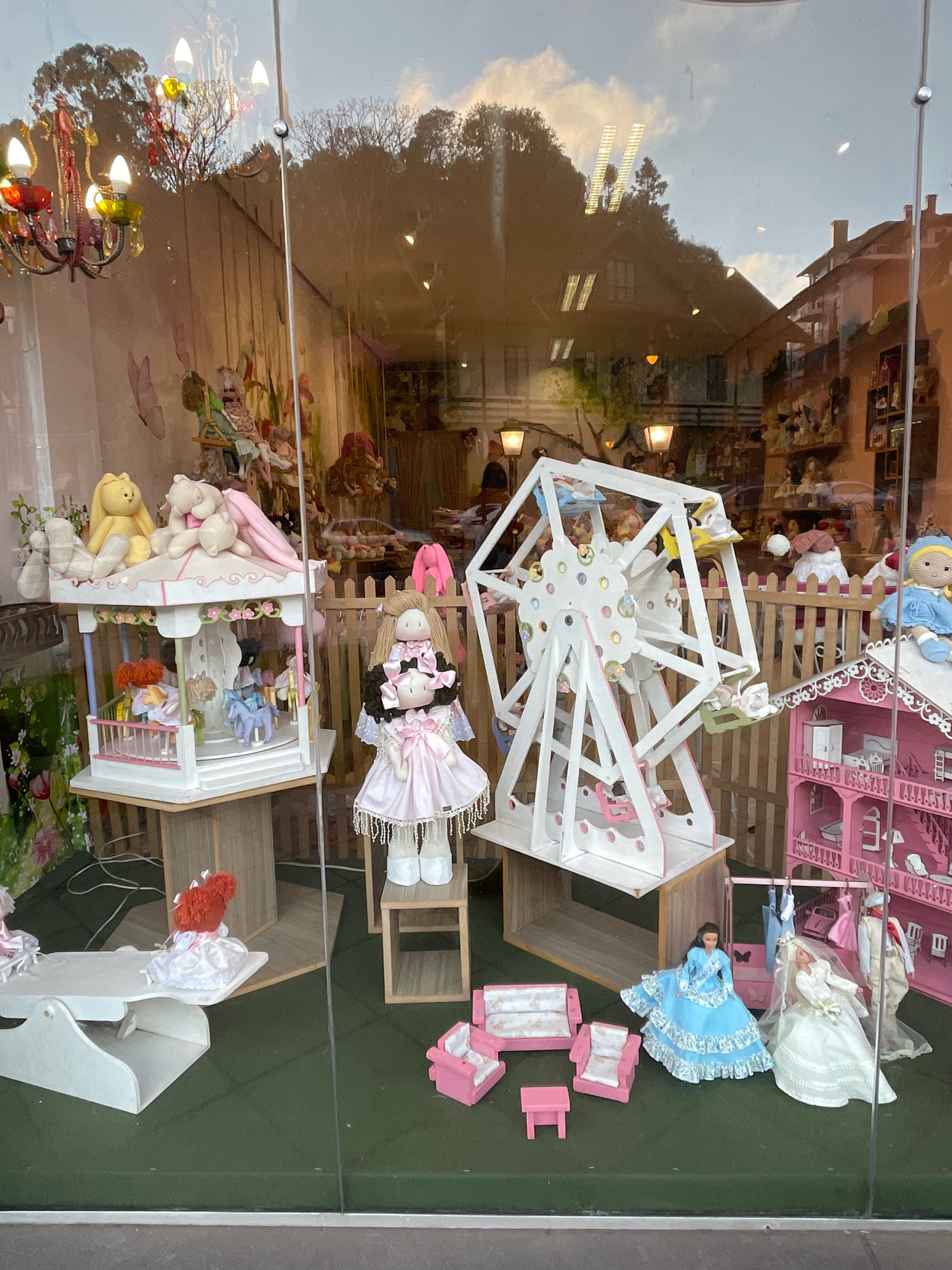 This is a photograph of a doll shop window display in Gramado, Brazil. The display features a variety of dolls and dollhouse accessories, arranged in a playful and whimsical scene. In the foreground, there are several pink miniature sofas and chairs, a small pink table, and a white miniature bed. To the left, a doll with bright red hair in a white dress sits on the bed. Further into the display, a large doll with blonde hair wears a pink dress and stands next to a miniature carousel. In the center of the display is a large white Ferris wheel with small dolls in each car. To the right, a pink dollhouse with multiple floors is visible, and next to it, two larger dolls are displayed: one in a blue dress and another in a white wedding dress. In the background, the reflection on the glass shows trees, buildings, and the sky. The overall impression is a charming and detailed doll display designed to attract customers.