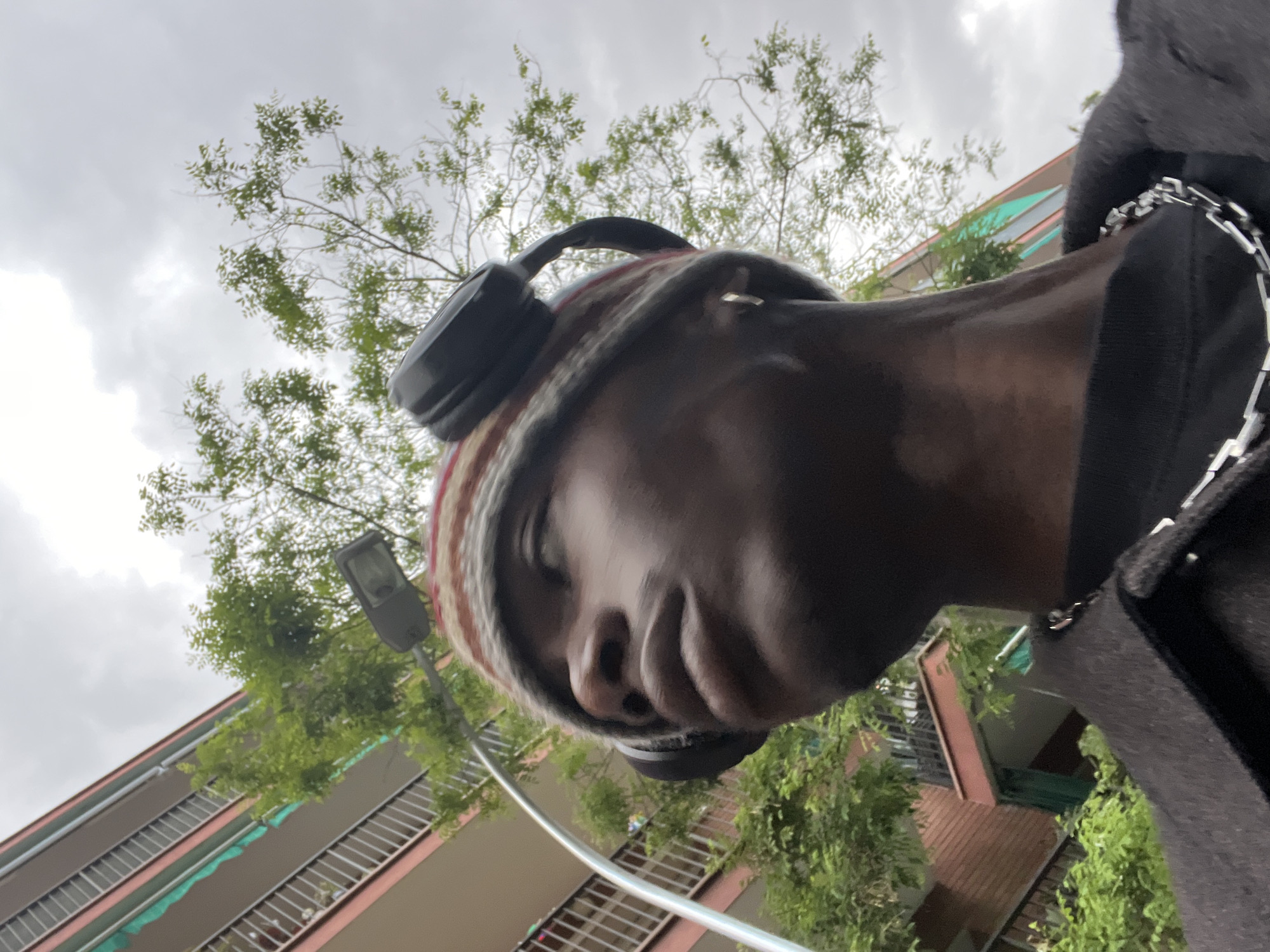The image shows a close-up, low-angle shot of a person wearing a striped beanie and headphones, facing downwards. The person has dark skin and appears to be male. Part of a black jacket or shirt is visible on the right side, adorned with a silver chain.
In the background, there's an apartment building with multiple floors, pink and green accents, and barred windows. Above the building are trees and an overcast sky. A street lamp is also visible amidst the foliage.
While identifying a precise location in Barcelona based solely on this image is difficult, the architecture and urban environment suggest a residential area within the city. It's impossible to identify the precise location in Barcelona from the image.