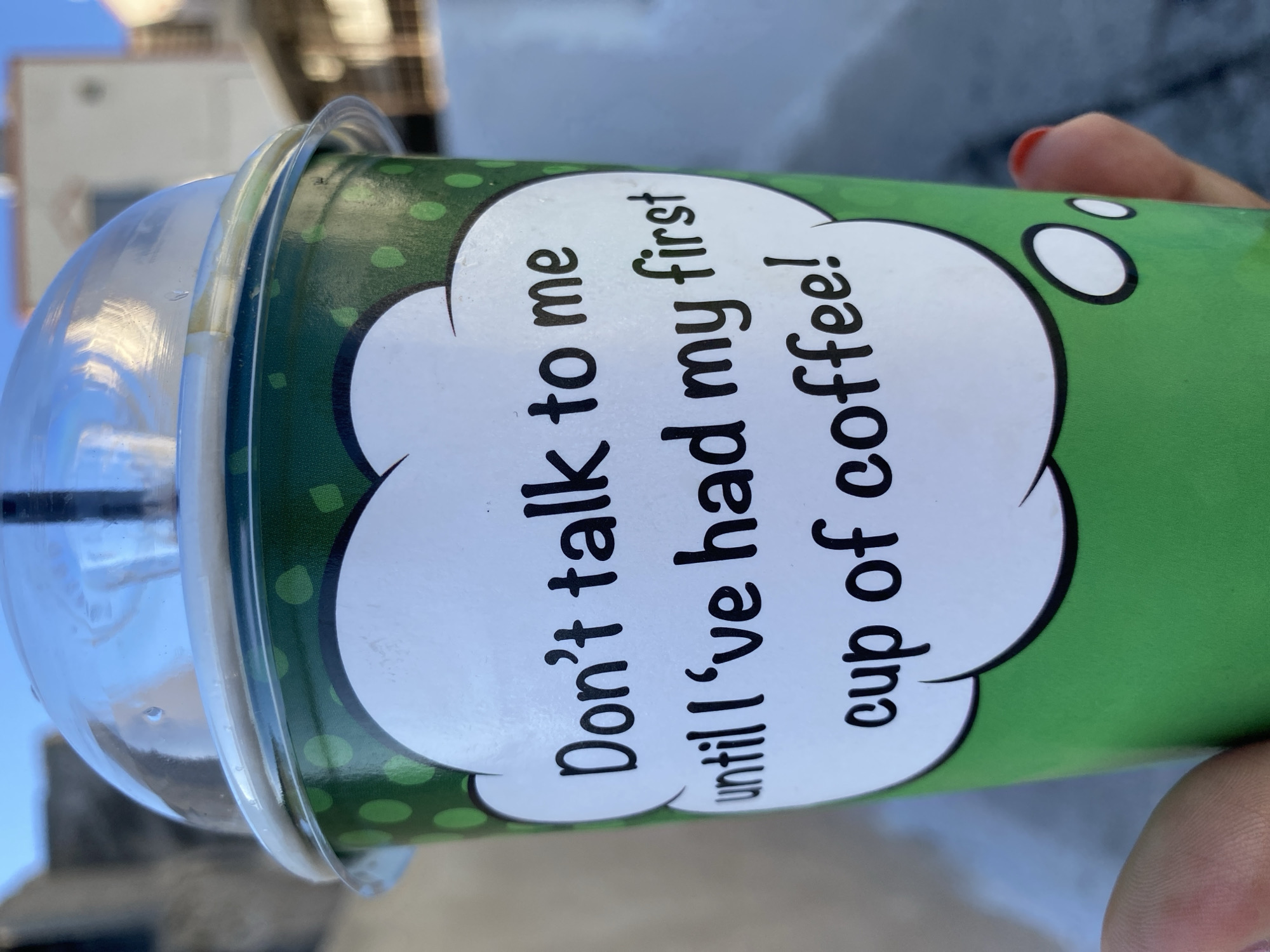 A close-up shot focuses on a green paper cup with white polka dots and a transparent plastic lid, holding a beverage with a black straw. The cup features a white, cloud-shaped graphic containing text that reads, "Don't talk to me until I've had my first cup of coffee!". The background shows out-of-focus buildings and a clear blue sky, suggesting an outdoor urban setting, possibly in Ia Municipal Unit, Greece. A person's hand with red-painted fingernails is visible on the right side of the frame, holding the cup. The lighting suggests it is daytime.