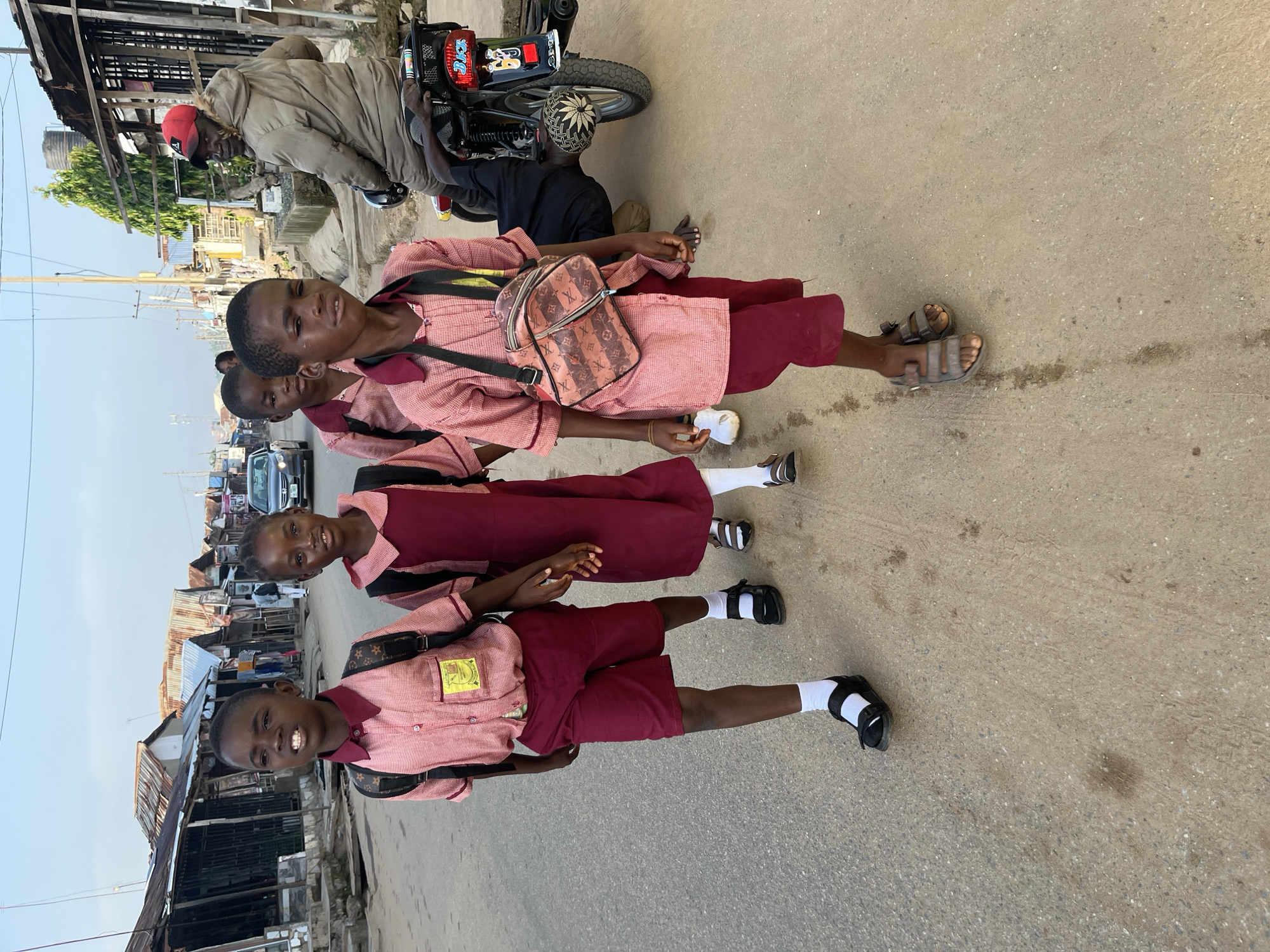 Four children in school uniforms walk down a dusty street in Abuja, Nigeria. The two children in the foreground, a boy and a girl, are wearing pink and burgundy checkered shirts with burgundy shorts and skirts, respectively. Both have black backpacks on. The boy is carrying a designer-style patterned bag over his shoulder. The two children behind them, a boy and a girl, are also in school uniforms. The street is lined with market stalls with corrugated iron roofs. A man wearing a beige jacket and a red cap is seated on a motorcycle to the right of the frame, facing away from the children. Cars and other vehicles are visible in the background, along with power lines and buildings. The sky is clear and the lighting suggests it is late afternoon or early morning.