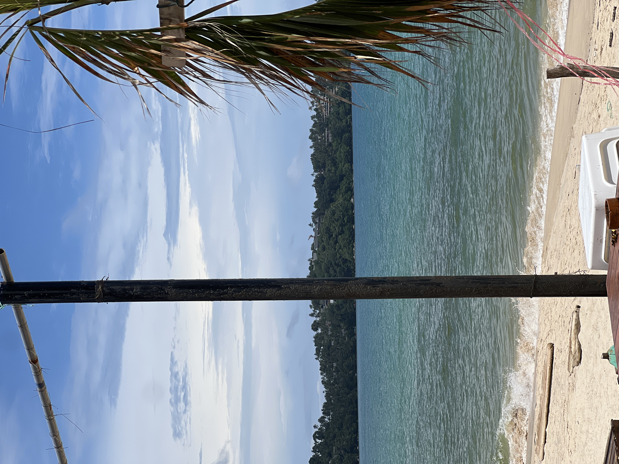 The image depicts a tranquil beach scene under a partly cloudy sky. The foreground is dominated by a dark, horizontal pipe or beam, with a lighter, bamboo-like structure running diagonally below it. A portion of a palm frond is visible at the top, framing the scene.
The middle ground features a sandy beach with a gentle wave lapping at the shore. A white beach chair is partially visible on the right side of the image, suggesting a place for relaxation. Beyond the beach, a lush green hillside covered in trees rises, with some buildings or structures nestled within the foliage. The sea is a calm, turquoise color, reflecting the bright sky.
The overall atmosphere is serene and inviting, typical of a tropical vacation spot. The time of day appears to be daytime, likely late morning or early afternoon, given the brightness of the sun and the clear shadows. The weather seems pleasant, with a mix of sun and clouds. There is no visible text in the image.