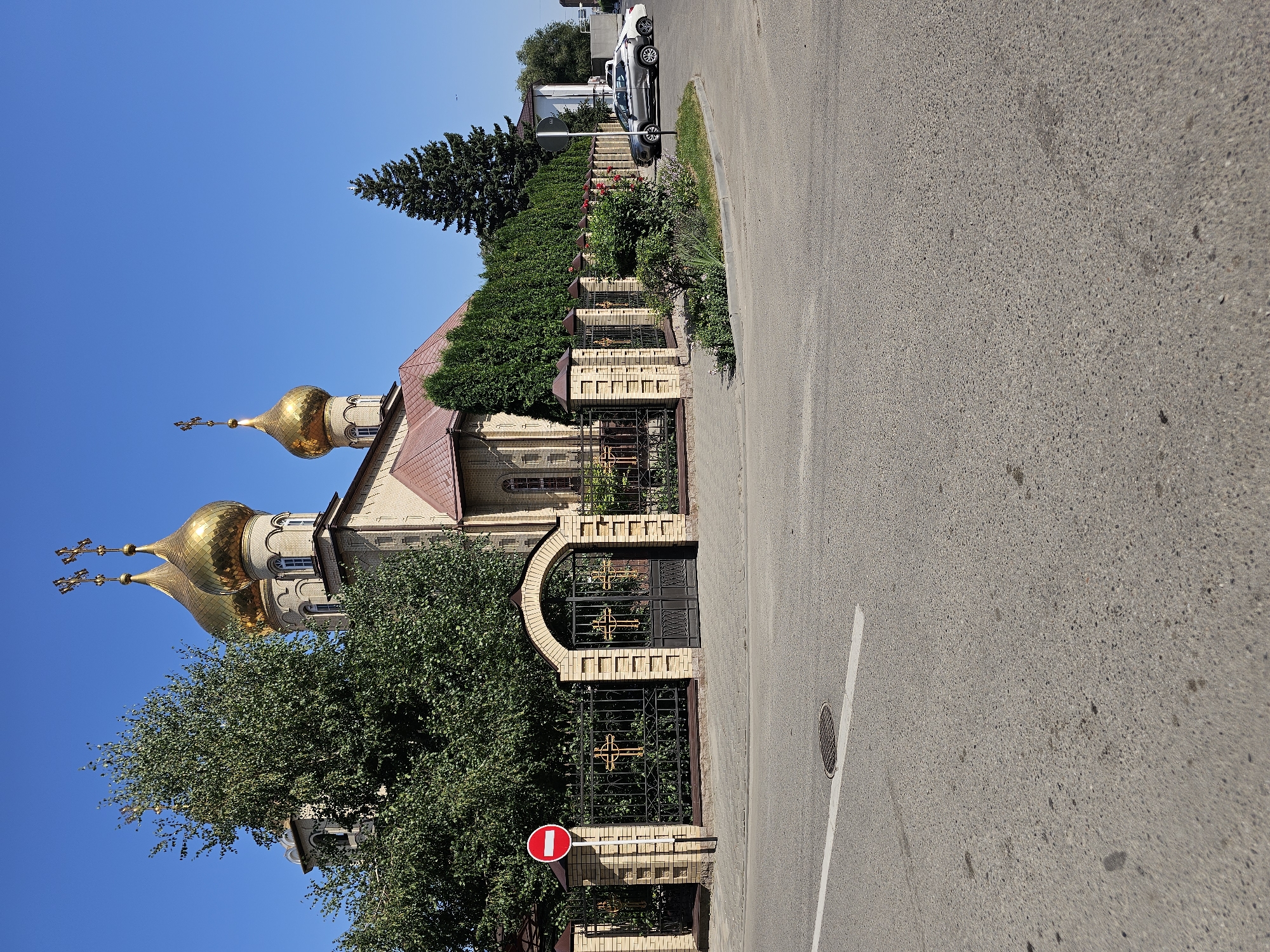 The image depicts the Church of the Archangel Michael in Cherkessk, Russia. The church features golden domes and is situated behind a brick fence with an arched gate. A "No Entry" sign is visible on the left side of the fence. The church is surrounded by trees and a dense hedge. In the foreground, a paved road with a white line and a manhole cover is visible. Two cars are parked on the side of the road near the church. The sky is clear and blue.