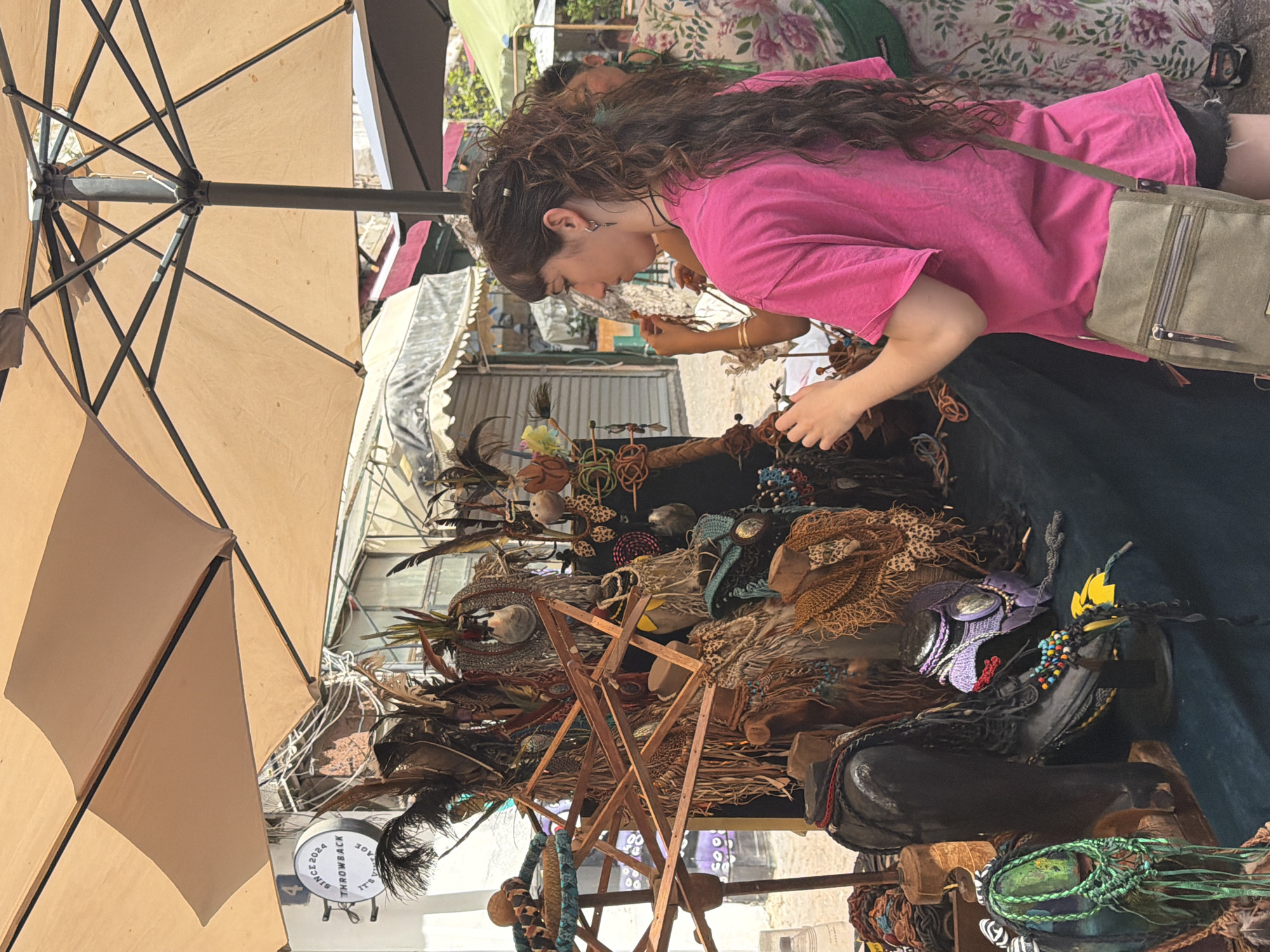 This media file captures a vibrant outdoor market scene, likely in Tel-Aviv, Israel, under bright daylight. The main subject is a young woman with long, dark, curly hair, wearing a bright pink t-shirt and a khaki-green cross-body bag. She is intently browsing a market stall, leaning over to inspect an array of handcrafted items.
The stall is laden with unique accessories and decorative pieces, many featuring feathers, beads, intricately woven materials, and leather. Some items appear to be elaborate headwear or masks, while others are necklaces or smaller ornaments. Prominently displayed are items resembling tribal or bohemian artistry. One distinct object is a black boot-like display piece adorned with colorful beads and various textures.
The scene is shaded by a large, beige market umbrella, which occupies the upper left and bottom left portions of the frame, its internal structure visible. In the background, other market stalls with fabric awnings and distant buildings can be seen, indicating a bustling outdoor environment. A circular white sign is partially visible in the lower left, with text that reads "SINCE 2024 THROWBACK IT'S A". Another person is partially visible behind the main subject, also seemingly engaged with the market. The woman's focused expression suggests she is deeply interested in the unique goods offered.