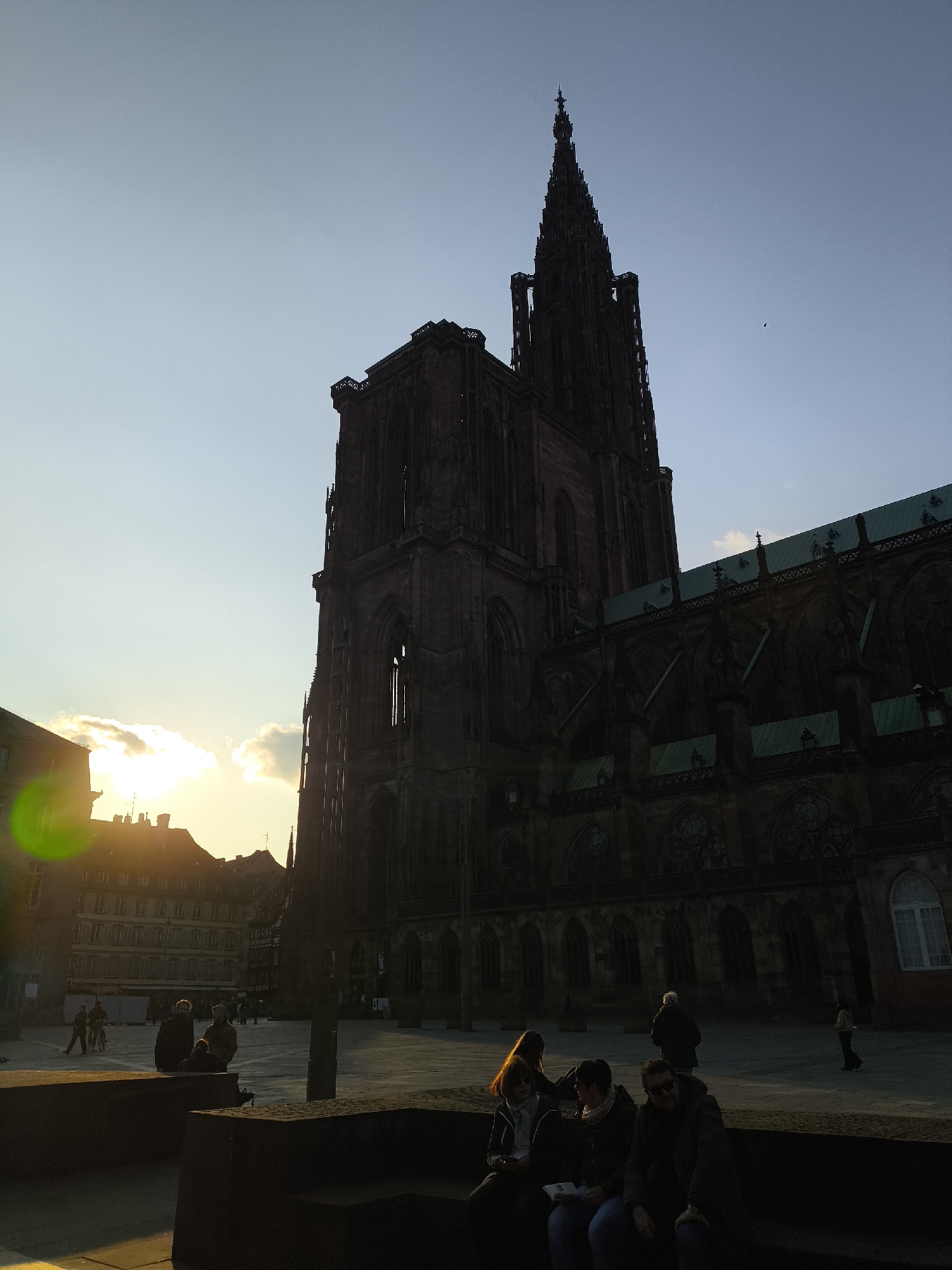 The image shows the Strasbourg Cathedral in France, a large Gothic structure, dominating the scene, with its intricate architectural details silhouetted against the evening sky. To the left, the sun sets behind the buildings, casting long shadows and creating a lens flare. In the foreground, there's a public square with a few people sitting on a low wall or benches. Additional people are walking around the square in the middle distance. The environment appears to be urban and historical.