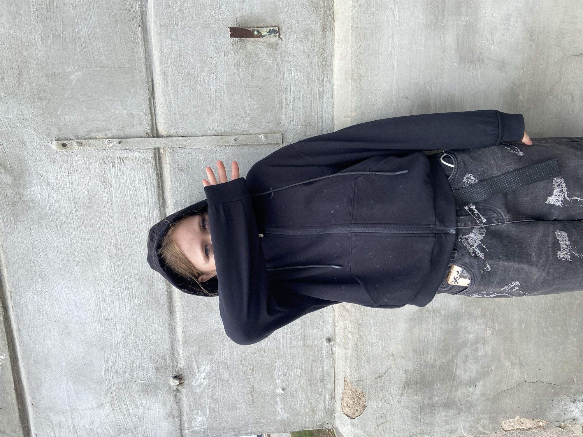 A person in a black hooded sweatshirt and gray jeans stands against a weathered concrete wall with metal bars. The person covers their face with a hand. The location is Votkinsk, Russia.