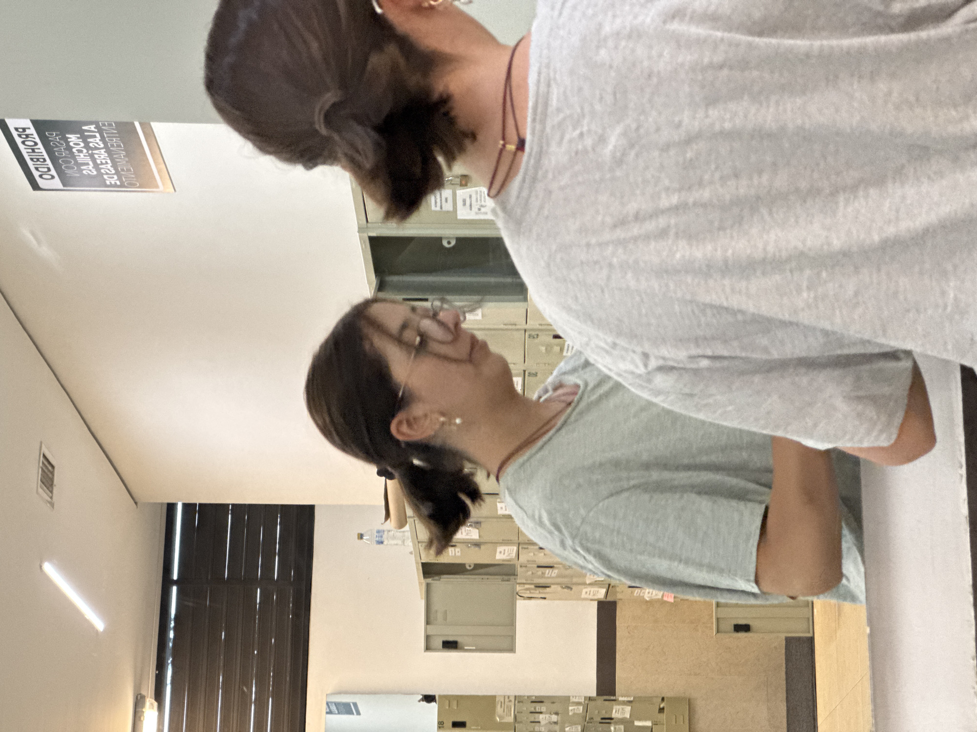 The image, rotated 90 degrees clockwise from its original vertical orientation, captures two individuals within an indoor setting, likely an office, archive, or storage room. In the foreground, an individual is seen from behind, wearing a light grey short-sleeved t-shirt and with dark hair tied in a bun at the back of the head. A reddish-brown necklace with small gold beads is visible around their neck.
Facing this person, in the middle ground, is another individual presented in profile, looking slightly upwards and to the right. This person wears a light blue-green t-shirt, glasses, and has dark hair pulled back into a low ponytail. A small pearl earring is visible on their ear. Both appear to be focused on or interacting with the objects in the background, suggesting they are accessing or organizing items in the storage units.
The background is dominated by numerous stacked beige metal filing cabinets or lockers, some with visible labels and one appearing partially open. A plastic water bottle rests on top of one of these cabinets. To the left, a window with vertical blinds allows some light into the room, and a fluorescent light fixture is visible on the ceiling/wall.
A white sign with dark text is mounted on the upper left wall. The text, reflected and partially mirrored, reads: "PROHIBIDO / COMIDA Y BEBIDA / LAS MOCHILAS / Y CARTERAS SE DEJARÁN / EN EL GUARDARROPA / MIENTRAS". This translates to: "PROHIBITED / FOOD AND DRINK / BACKPACKS / AND WALLETS WILL BE LEFT / IN THE CLOAKROOM / WHILE". The scene suggests an administrative or functional space, consistent with the provided location context of San Andrés Cholula, Mexico. The time of day is not discernible, but the setting is clearly indoors.