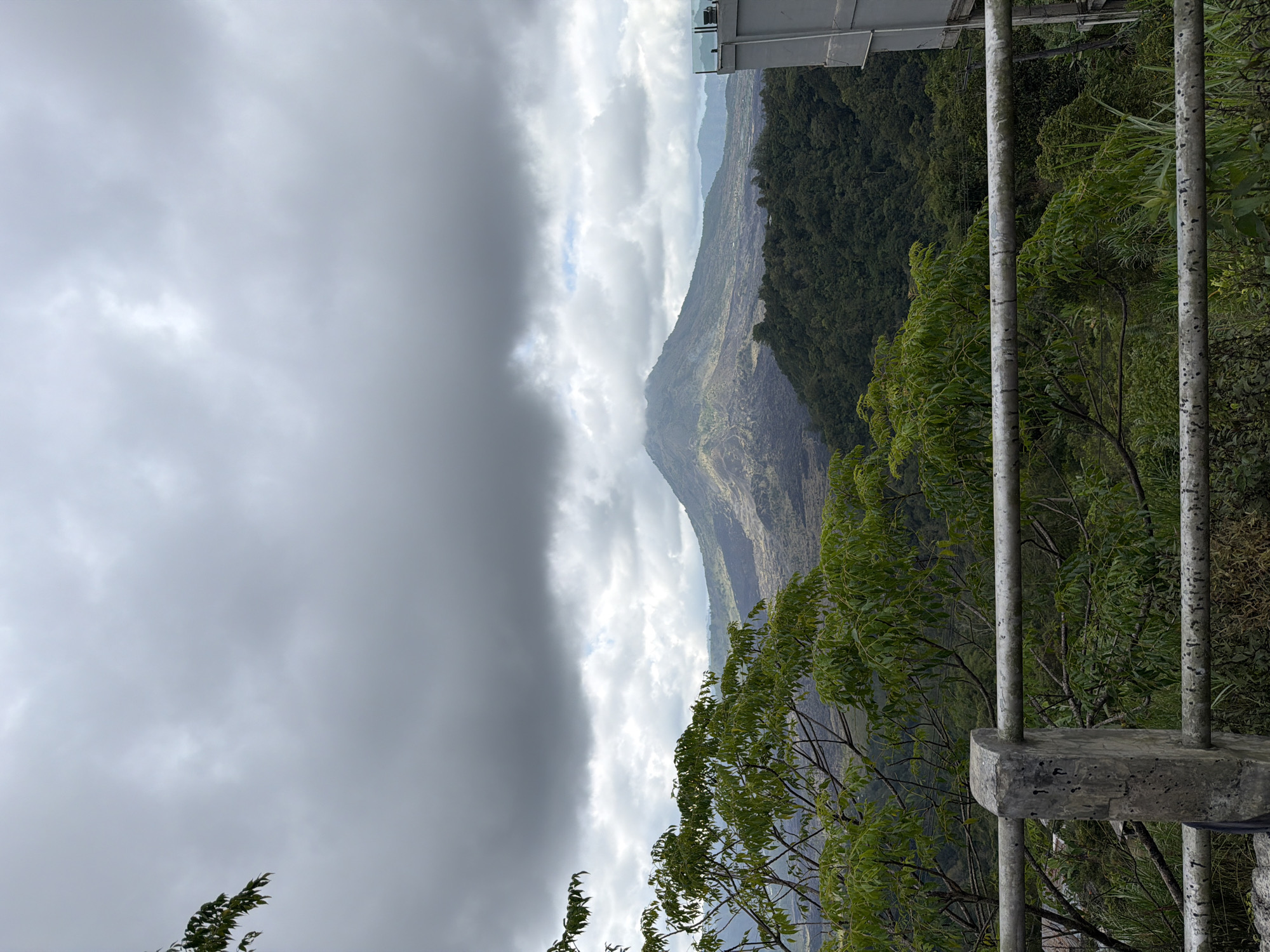 The image captures a panoramic view of Mount Batur in Kintamani, Indonesia, under a cloudy sky. In the foreground, a weathered concrete railing with metal bars runs horizontally across the frame, partially obscuring a lush green canopy of trees and foliage. Beyond the railing, the rugged, dark slopes of Mount Batur dominate the mid-ground, with patches of lighter vegetation visible on its sides. The base of the mountain is covered in a dense forest. In the far right, a modern building with glass railings is partially visible, suggesting a viewpoint or a nearby structure. The sky is overcast with heavy grey clouds, with a few brighter patches indicating diffused sunlight. The overall scene conveys a sense of natural grandeur and a somewhat somber atmosphere due to the cloudy weather.
