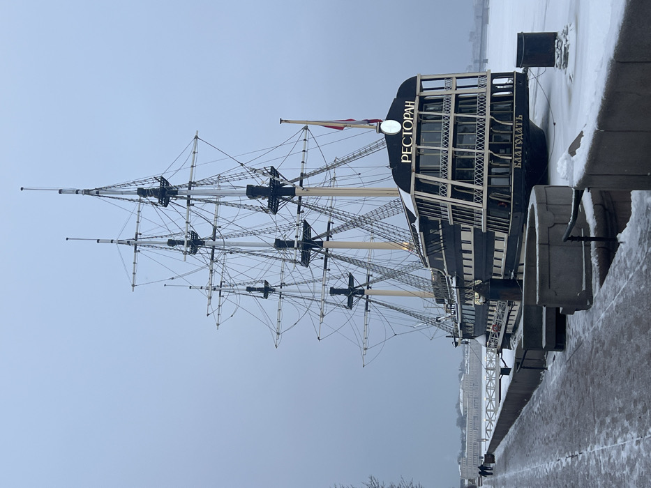 The image depicts the Blagodat' (БЛАГОДАТЬ) restaurant ship, a multi-masted sailing vessel, moored on the Petrovskaya Embankment of the Bolshaya Neva River in Saint Petersburg, Russia, during winter. The scene is characterized by a uniformly overcast, light gray sky and widespread snow cover.
The ship itself has a dark hull, possibly black or dark blue, with numerous light-colored rectangular windows arranged across multiple decks. The stern features a prominent, multi-story glassed-in structure, typical of a restaurant, with the word "РЕСТОРАН" (Restaurant) illuminated in gold lettering above it. Below, the ship's name "БЛАГОДАТЬ" is also displayed in gold. Four tall masts with intricate rigging rise from the deck, extending into the sky. A red and yellow flag flies from a pole at the ship's stern.
The frozen river adjacent to the ship is completely covered in a layer of white snow. The ship is moored to a concrete embankment, parts of which are also snow-covered. A grey paved walkway runs along the embankment, showing patches of snow alongside cleared sections. Concrete barriers, topped with snow, line the edge of the embankment overlooking the frozen river.
In the mid-ground to the left, two indistinct figures are walking on the snow-dusted paved path. In the far distance across the frozen river, a long, light-colored historical building complex, potentially featuring a colonnade, is visible against the muted horizon. Further to the right in the background, a faint outline of a bridge can be discerned. The overall environment is cold, tranquil, and urban.