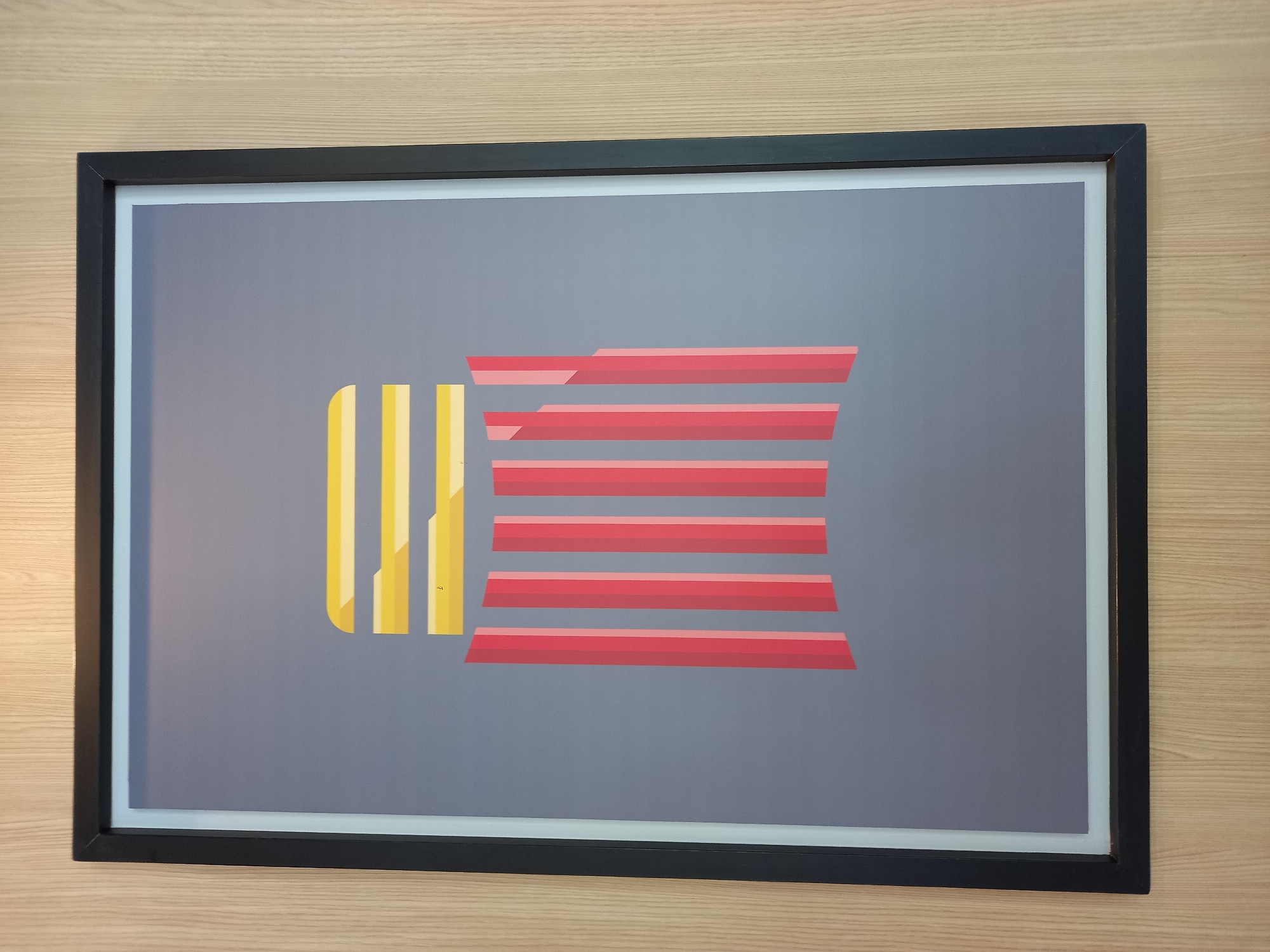 A framed abstract art piece hangs on a light-colored wood-paneled wall. The artwork features a grey background with a stylized illustration of a hamburger. The top of the "bun" is represented by three horizontal yellow bars, and the "patty" and "bun" are depicted by several vertical red bars of varying heights, creating a layered effect. The frame is black.