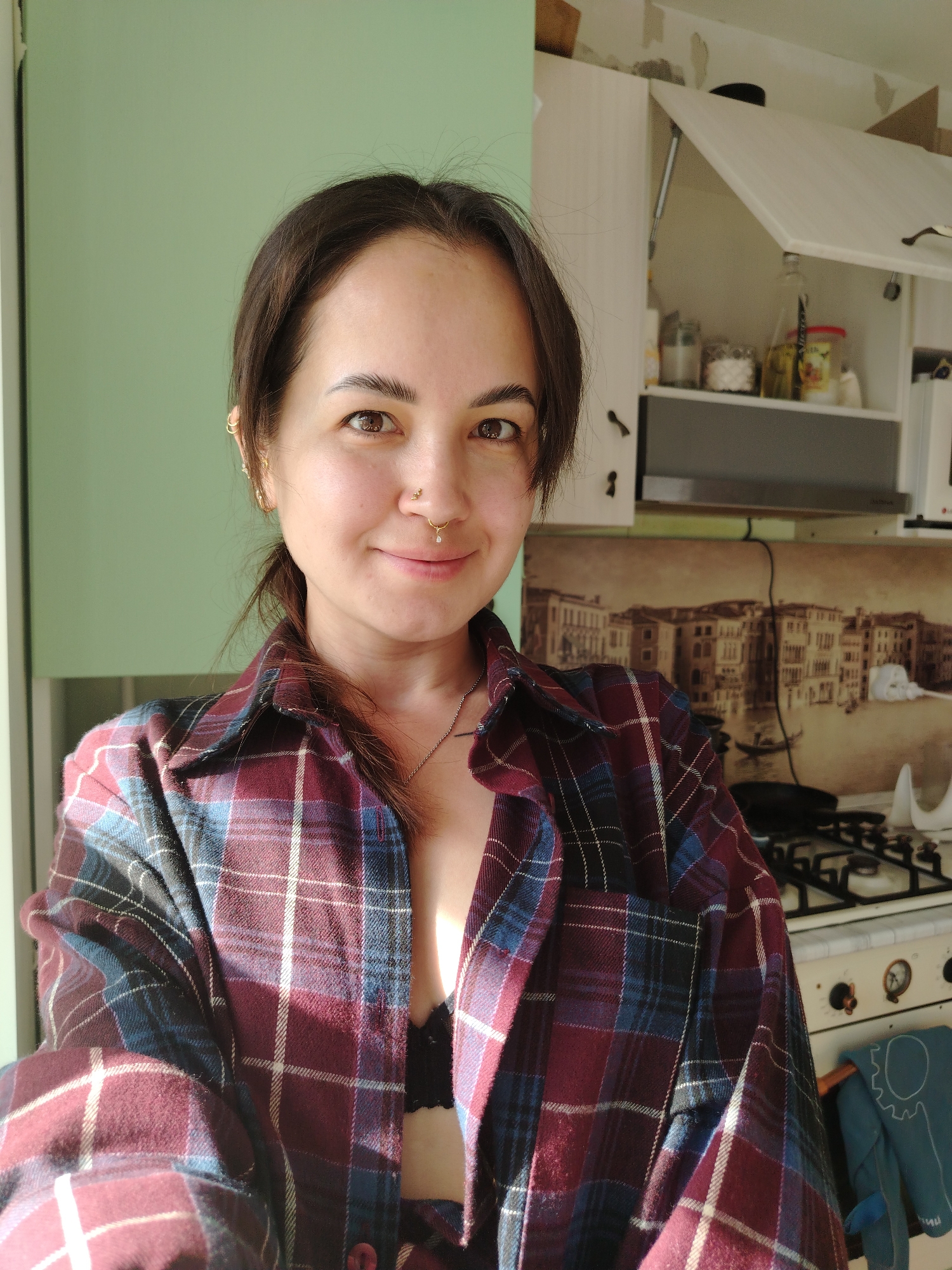 A medium shot depicts a woman indoors, likely a kitchen, wearing an unbuttoned, plaid flannel shirt over a black bra. She has brown hair pulled back, multiple piercings in her left ear, and a nose piercing. Behind her is a light green wall on the left and kitchen cabinetry above a stove on the right. A decorative panel showing a cityscape of Venice, Italy is behind the stove. The gas stove is white and vintage looking. An oven mitt hangs from the oven door handle. Noviy, Russia is not visible in the image.