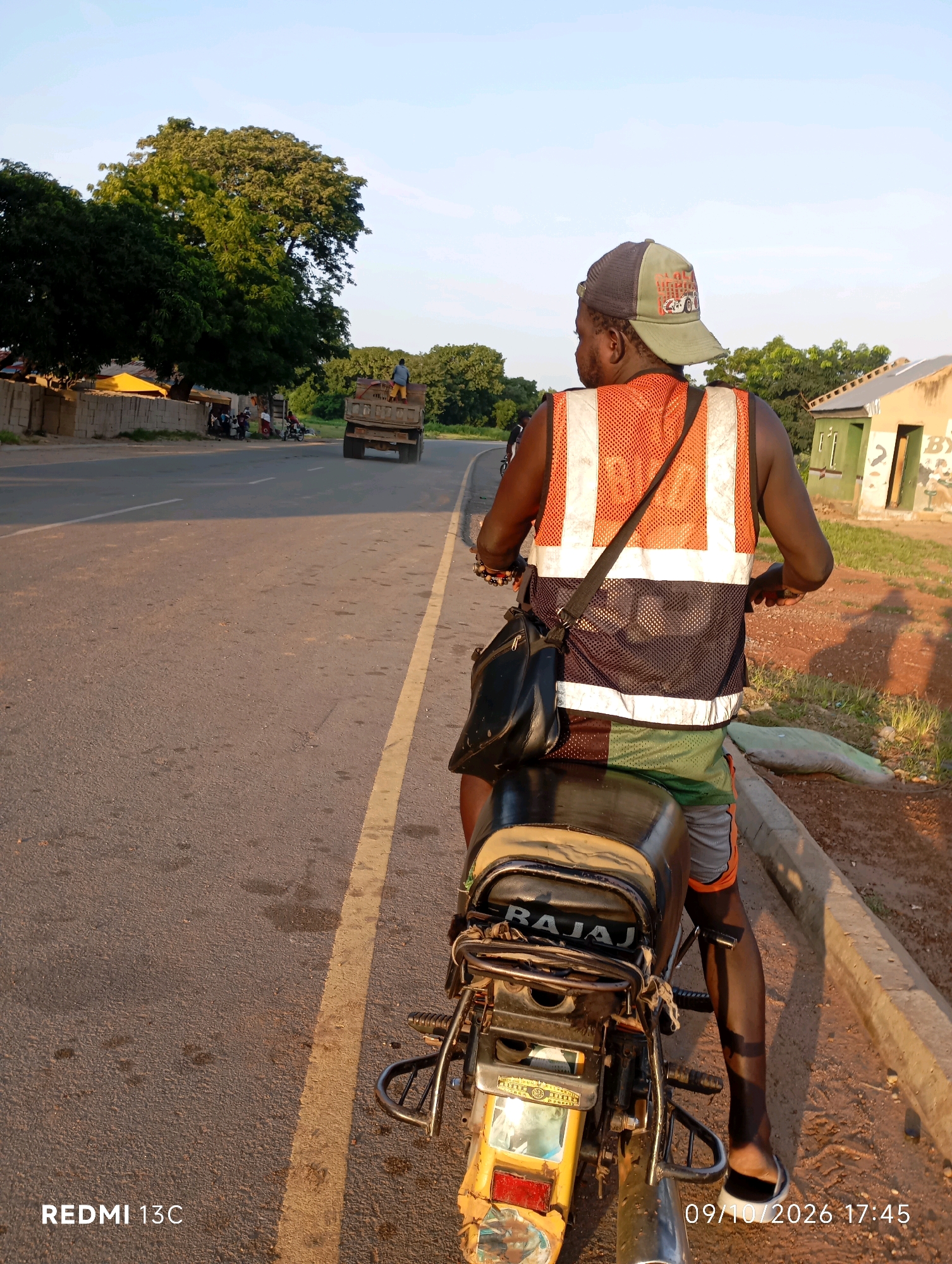 In Shendam, Nigeria, a person wearing a high-visibility vest and a baseball cap rides a motorcycle. The rider is seen from behind, with the motorcycle's rear end prominently in the foreground. The motorcycle is on a paved road with a yellow line marking the lane. In the background, a truck is visible, and on the side of the road, there are buildings and trees. The sky is clear and the lighting suggests it is either early morning or late afternoon. The overall scene depicts a moment of transit within a semi-urban or rural setting in Nigeria.