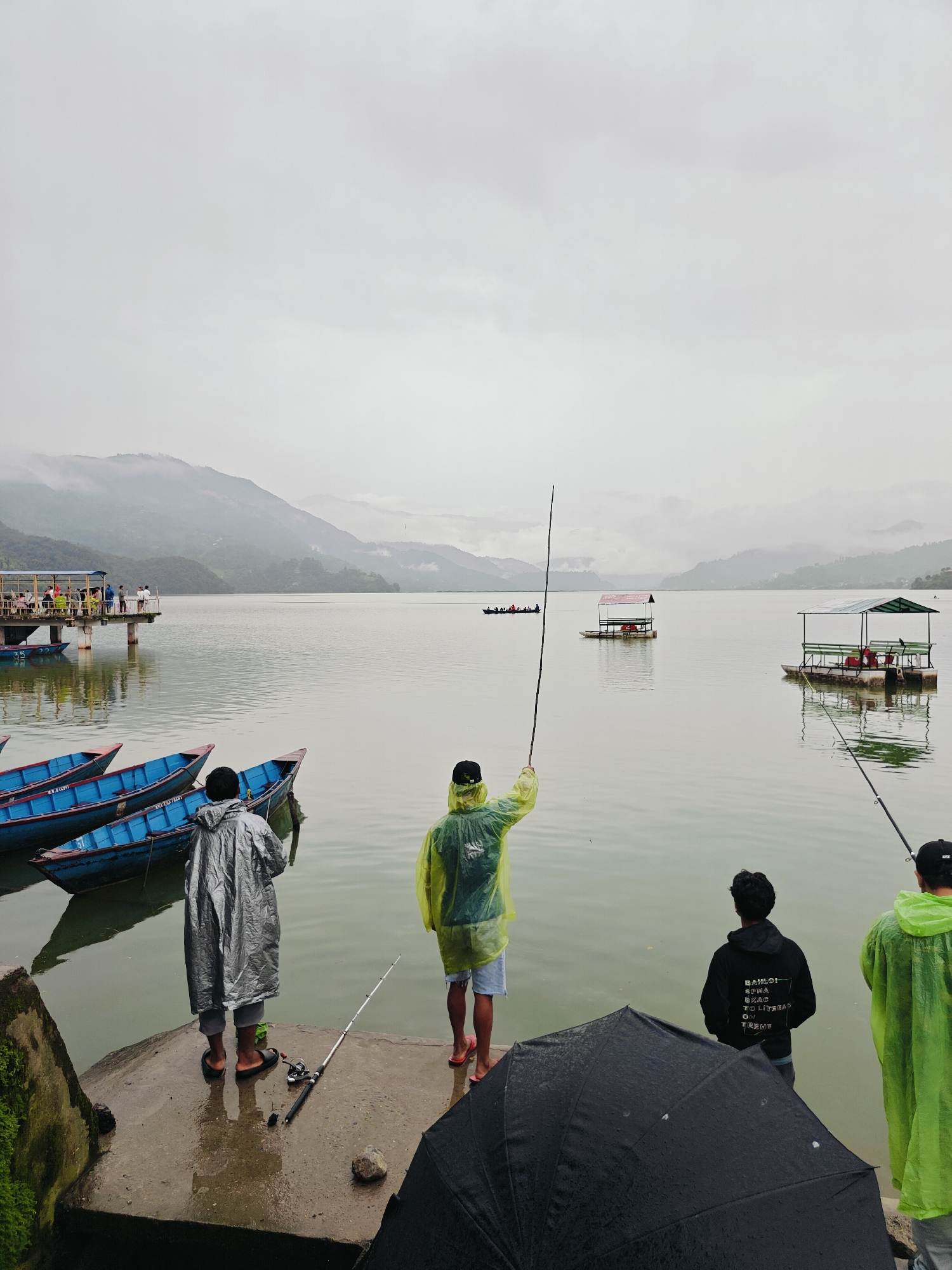 This is a photograph taken on a cloudy day at Phewa Lake in Pokhara, Nepal. The image captures a group of people engaged in fishing activities. In the foreground, four individuals are standing on a wet, concrete platform. Two are wearing brightly colored raincoats, one yellow and one bright green. The person in the yellow raincoat is holding a fishing rod and facing the lake. Another person, wearing a grey raincoat, is standing with their back to the camera, also facing the water, with a fishing rod and reel at their feet. A fourth person, wearing a black hoodie, stands between the two in raincoats, looking out at the lake. A partially open black umbrella is in the lower right corner.
To the left of the foreground, several blue boats are docked along the edge of the platform. In the mid-ground, a pier extends into the lake with a group of people on it. Further out on the lake, two floating structures with roofs are visible, possibly serving as recreational or resting spots. A boat with several people on board is also visible in the distance. The background is dominated by the misty outlines of the Annapurna mountain range, typical of Pokhara's scenic landscape. The overall atmosphere is serene, with the overcast sky casting a soft light over the tranquil waters of Phewa Lake.