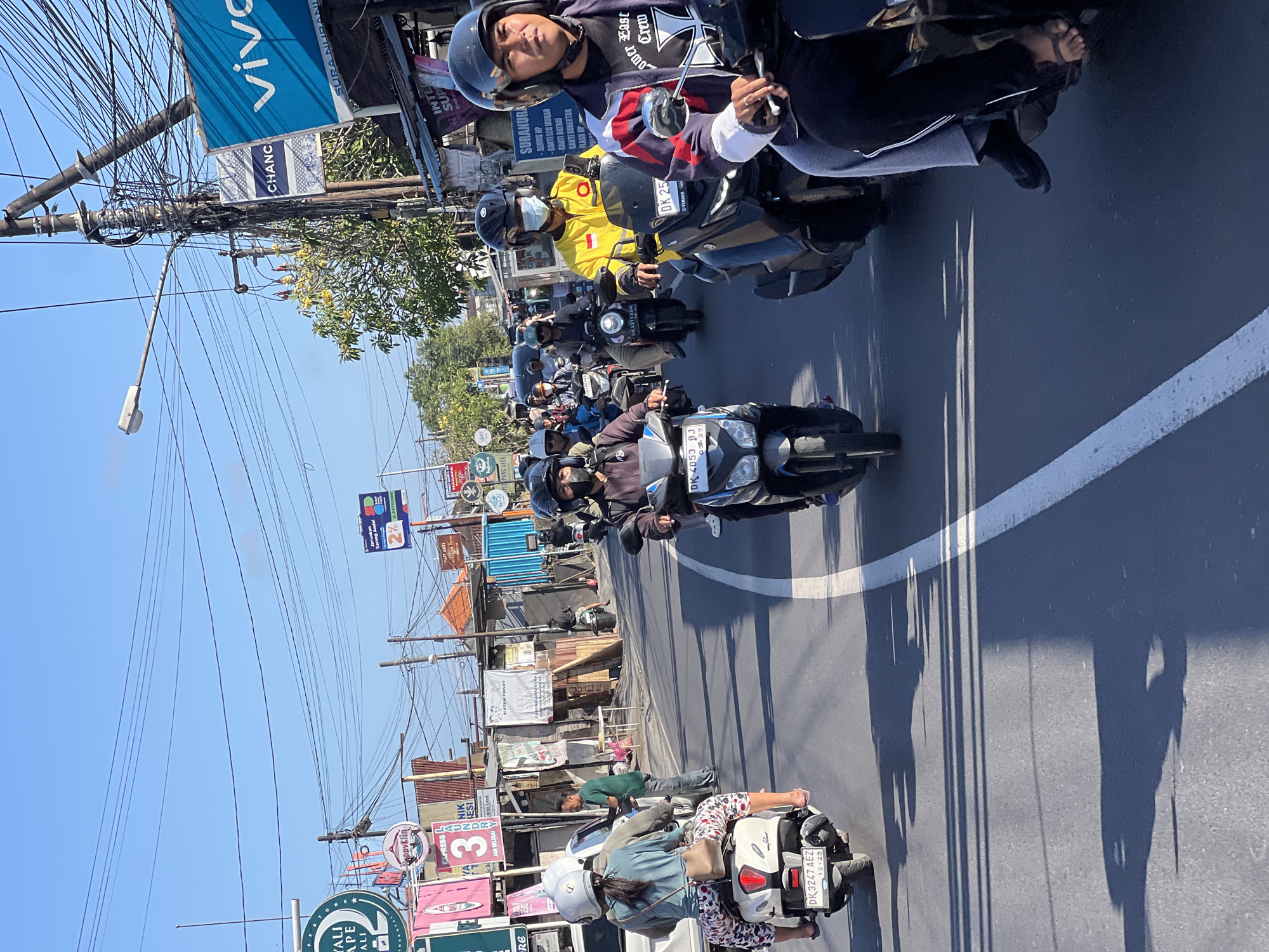 This is a street-level, eye-level, medium shot captured on a bright, clear day in Denpasar, Indonesia, depicting traffic congestion. The scene is dominated by a procession of motorbikes traveling down a paved road.
In the foreground, the right side of the frame features a rider wearing a black and white jacket with red accents, a black helmet, and a black shirt. They are on a dark-colored motorbike, partially obscured. To their left, a motorbike with a black front and silver accents is visible, ridden by a person wearing a black helmet and a mask. The number plate reads "DK 4053 BJ".
Further back, a motorbike with a rider in a yellow jacket and black helmet is prominent. This rider's motorbike has a number plate "DK 25...". To their left, a motorbike with a dark front is ridden by someone in a black helmet and a black shirt. The number plate reads "DK 5371 AGW".
On the left side of the road, a scooter with a white body is occupied by two people. The rear passenger, a woman, wears a light blue shirt and patterned trousers, with a light-colored helmet. The front rider, also in a light-colored helmet, is out of frame. The scooter's number plate reads "DK 3247 AEZ".
The surrounding environment includes a variety of signs and poles lining the street. Notable signs include a blue "vivo" advertisement, a "CHANC" sign, and various other advertisements and shop signs in Indonesian. Buildings with corrugated metal roofs are visible behind the shops. Power lines and streetlights crisscross the sky. The road has a white dividing line. The overall impression is of a busy urban street scene with moderate vehicular traffic.
