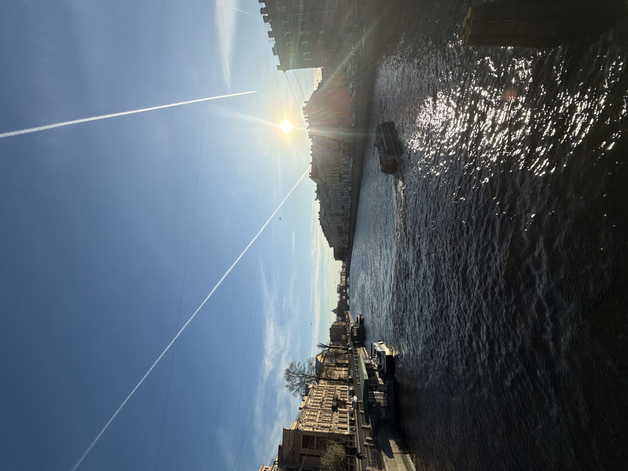 The image depicts a canal in Saint Petersburg, Russia, with buildings lining its banks. The sky is clear blue with visible contrails. Sunlight glints off the water's surface. Boats are docked along the canal. The photo appears to have been taken from a high vantage point. The sun is high in the sky and slightly obscured by the buildings, which are historical and typical of the city's architecture.