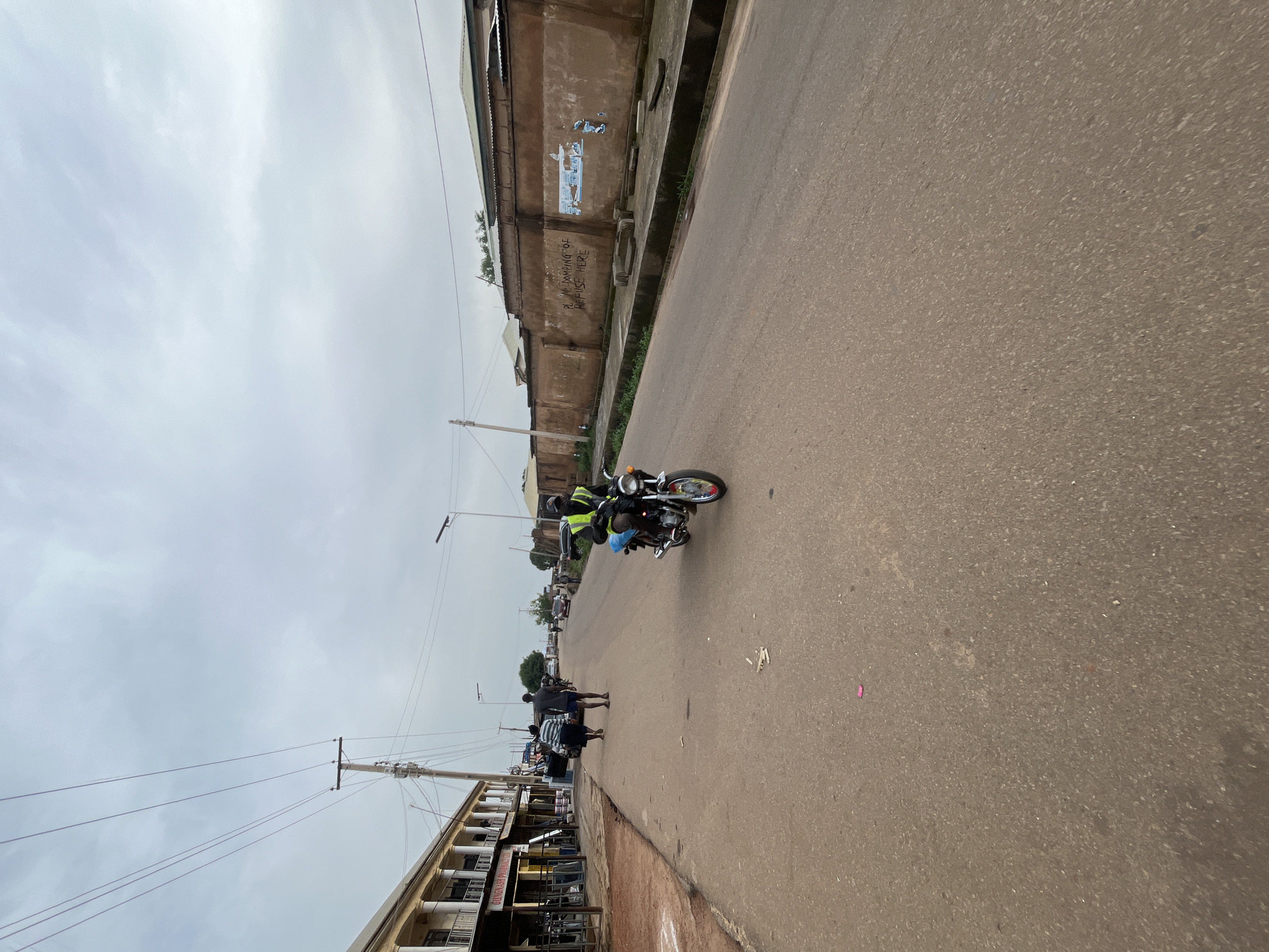 The media file shows a street scene in Gura Top, Nigeria. The perspective is from a low angle, looking upwards and sideways, with the horizon tilted. The sky is overcast with pale clouds.
The main subjects are people and a motorcycle on a paved road. A person wearing a neon yellow vest is riding a motorcycle. In the background, there are a few other individuals walking or standing by the roadside. The setting appears to be a commercial or residential area with buildings lining one side of the street. These buildings have a rustic appearance, with corrugated metal roofs and weathered walls. One building has visible graffiti or painted text that reads, "PL. M. DAMPING OF REFUSE HERE."
The scene depicts everyday street activity, likely during the day given the ambient light. The weather seems to be cloudy or overcast, suggesting it might not be a sunny day. The overall atmosphere appears calm and ordinary, with no indication of any special event. The presence of utility poles with wires and the style of the buildings provide clues about the location in Nigeria. The road surface is asphalt and appears to be in moderate condition. Some scattered debris is visible on the road.