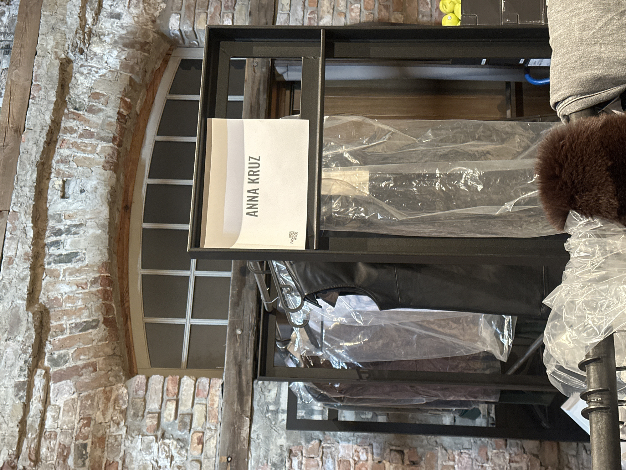 This image captures an indoor scene within what appears to be a historic or industrial building, characterized by rough, exposed brick walls and large, aged wooden beams. A prominent feature of the background is a tall, arched window with white frames and multiple panes.
In the foreground, a black metal shelving unit or clothes rack is visible, filled with garments. Many of these garments are protected by clear plastic garment bags. On the top right shelf, a grey sweater or hoodie is present, alongside a dark brown, fluffy item that could be fur. A black leather-like garment hangs lower down.
A notable object is a white sign or document placed on one of the shelves, clearly displaying the text "ANNA KRUZ" in black uppercase letters. Below this name, a small, partially obscured logo can be seen, which appears to include the words "FASHION WEEK". There are also some small yellow items on an upper shelf and a blue cable visible beneath a lower shelf.
No people are present in the image. The setting, with its architectural details, garments, and the "ANNA KRUZ" sign potentially linked to "FASHION WEEK," strongly suggests a context related to fashion, such as a showroom, boutique, or a backstage preparation area for an event, located in Riga, Latvia. The time of day and weather are not discernible.