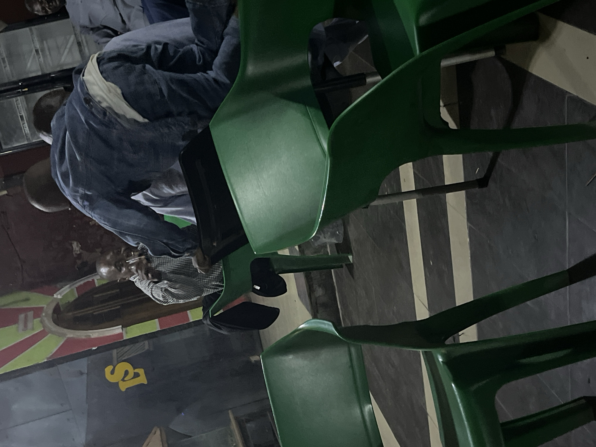 The image captures a dimly lit indoor scene, possibly a bar or social gathering space, in Jos, Nigeria. The foreground is dominated by the backs of several vibrant green plastic chairs. In the mid-ground, a man in a checkered shirt is seated, holding a glass to his mouth, appearing to drink. Behind him, a person wearing a denim jacket and jeans is leaning forward, their back to the camera. Another individual, wearing a light blue shirt, is visible to the right. The background features a curved architectural element with red and green striped decoration, and a yellow "LS" sign is affixed to a dark wall. The flooring is tiled with a dark and light grey checkered pattern.