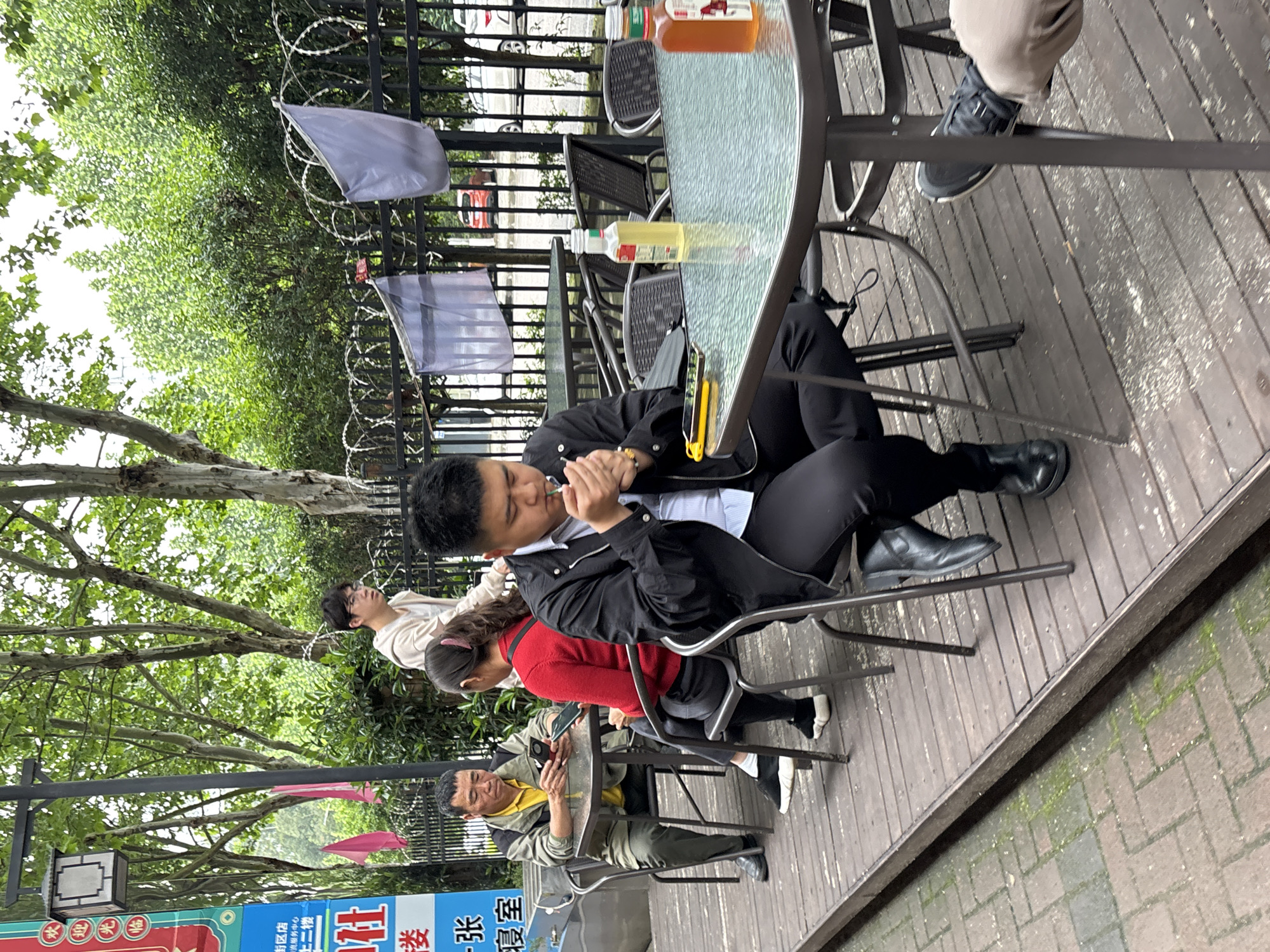 The image shows an outdoor seating area in Jiangjin, China. A group of four people sit at a round, glass-topped table on a wooden deck. The man closest to the camera wears a black jacket and pants, with a white shirt underneath, holding an object to his mouth. A woman in a red sweater sits next to him, and an older man in a yellow shirt and green jacket looks at his phone. A fourth person is visible behind them. The table holds two bottles of liquid. In the background, a black metal fence with barbed wire and white flags borders the area. Trees and greenery surround the space. Part of a colorful sign is visible at the bottom left corner of the image.