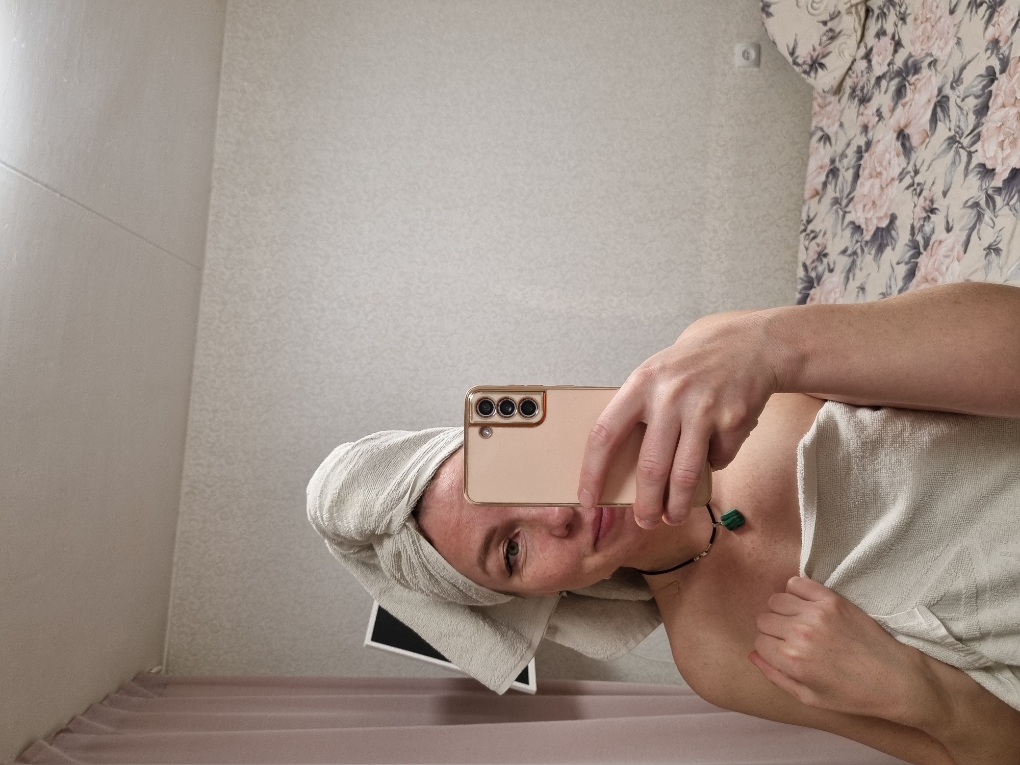 An indoor mirror selfie captures a person, likely female, from the chest up. She has a light-colored towel wrapped around her head and another towel draped around her body, covering her bare shoulders and chest. Her face, seen partially, has a neutral expression as she looks towards the gold-colored smartphone she is holding to take the picture. A dark cord necklace with a prominent green, possibly malachite, pendant hangs around her neck.
The background shows two different wall coverings. One wall features a subtle, light-colored patterned wallpaper. Another section displays a floral pattern with large pink and grey flowers, where a white electrical outlet is also visible. A textured, light-pink fabric, possibly a curtain or bedspread, is visible in the lower part of the scene. Behind her head, reflected in the mirror, is a dark, rectangular object, appearing to be a tablet or laptop screen. The setting suggests a residential interior, potentially a bathroom or bedroom, located in Chaikovsky, Russia. No visible text is present.