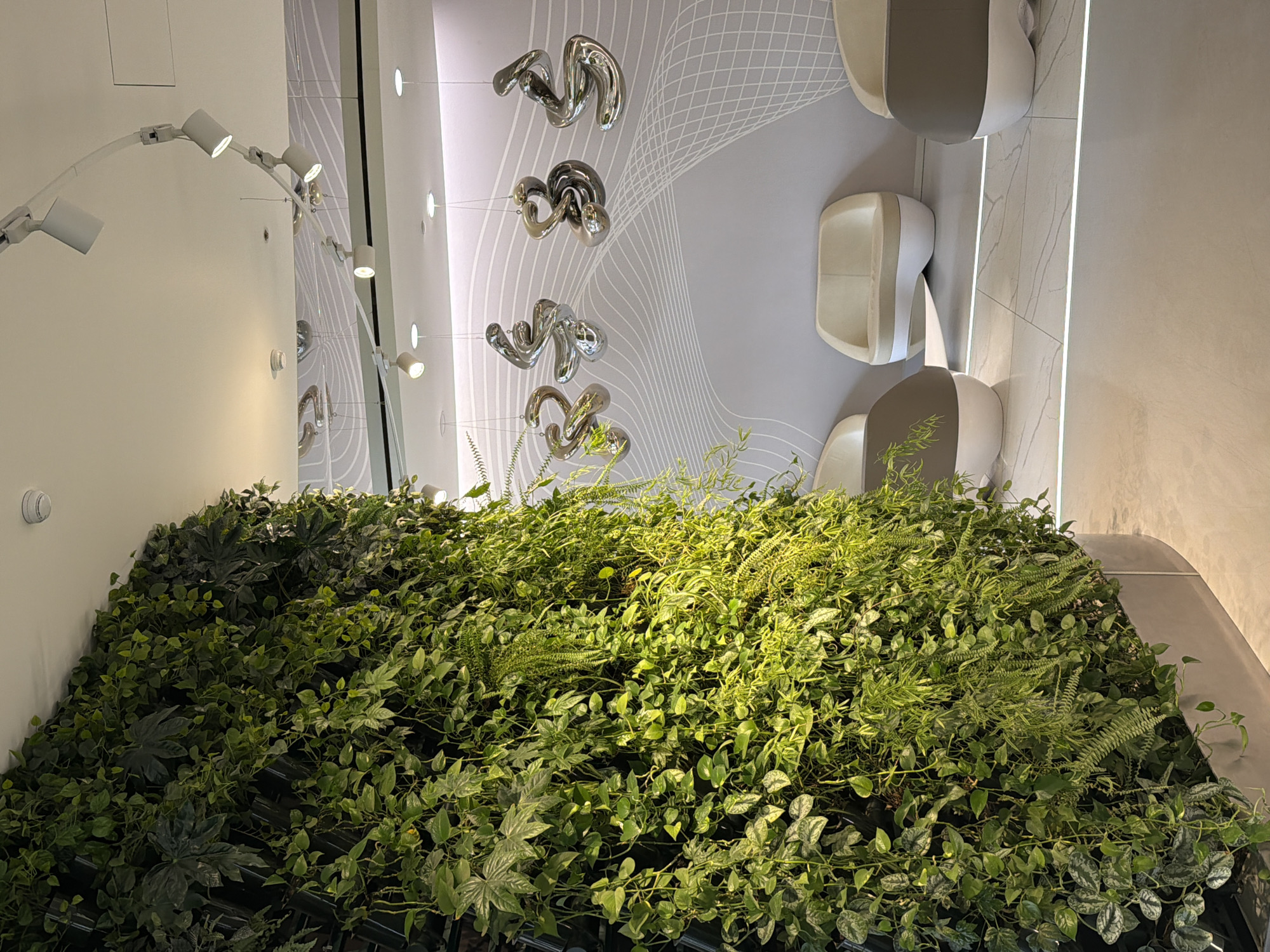 The image shows an indoor scene, likely a public restroom in Moscow, Russia. A wall of greenery covers the lower portion of the frame, creating a "living wall" effect. Above the greenery are modern, stylized urinals mounted on a grey-toned wall with decorative wave patterns. Chrome sculptures hang in the space above the urinals. A curved lighting fixture with multiple spotlights is mounted on a wall to the left, which also appears to reflect the interior. The overall aesthetic is contemporary, clean, and incorporates elements of nature.