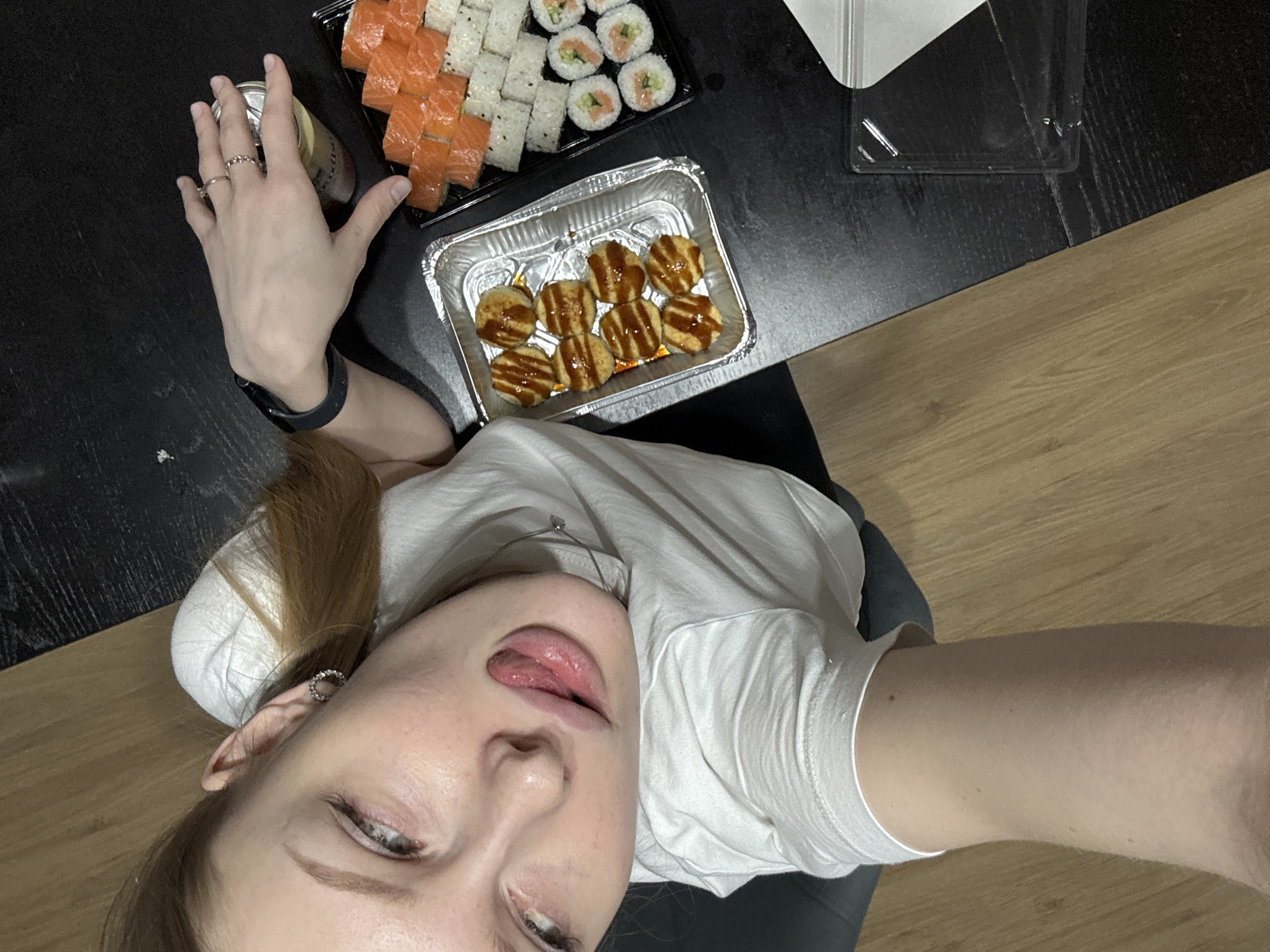 A high-angle indoor photograph captures a young woman from the chest up, positioned alongside a table laden with food. She is looking upwards towards the camera, with her head slightly tilted. Her mouth is slightly open, revealing her tongue in a relaxed or playful expression. She has light-colored hair and wears a plain white t-shirt. Her left hand, adorned with rings on her fingers and a dark smartwatch on her wrist, holds a light-colored beverage can.
On a dark, textured table surface beside her, a spread of food is visible. Two black rectangular trays contain various sushi rolls; one features numerous salmon-topped rolls, while the other holds white-rice rolls, some with visible fillings like salmon and cucumber, and smaller maki rolls. A silver foil container sits next to the sushi, holding several round, breaded or fried items drizzled with a dark, glossy sauce. A clear plastic container or lid is also present on the table. The table abuts a light-colored wooden floor, suggesting a casual home or dining environment. The overall scene depicts a relaxed meal or snack time, likely a selfie or personal photograph given the angle and pose. No specific time of day or location details for Perm, Russia are discernible from the visual cues, and no readable text appears on any of the objects.