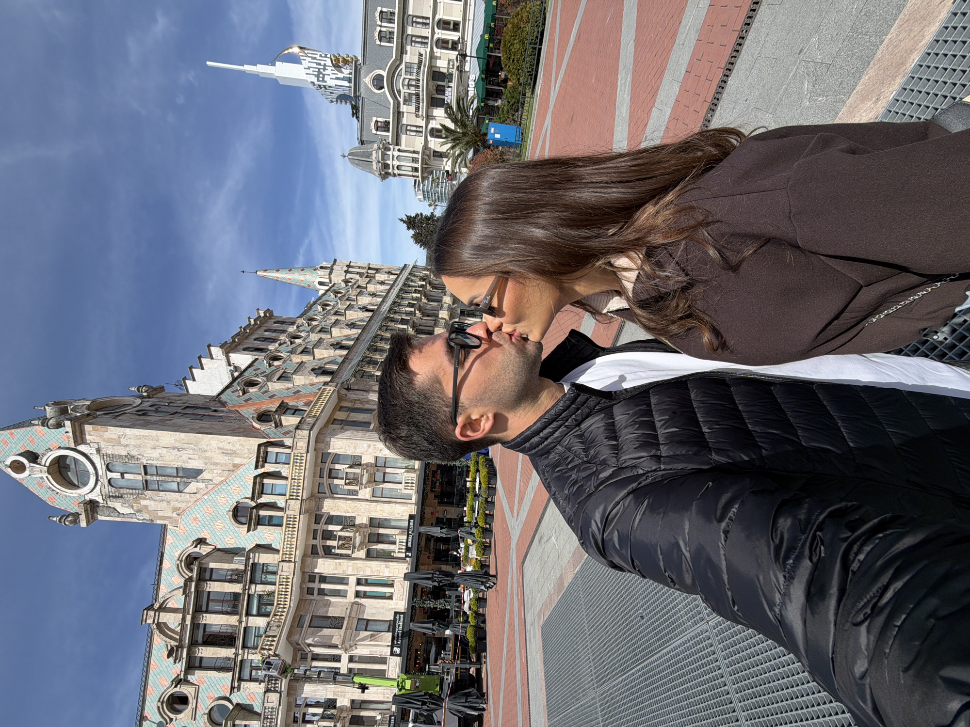 In Batumi, Georgia, a couple kisses in front of Europe Square. The man wears a black puffer jacket and dark sunglasses. The woman wears a brown coat and sunglasses. Two buildings stand behind them: one with a decorative green and blue tile roof, and another with a white spire. The square is paved with red and grey bricks and has metal grates. Outdoor seating for restaurants is visible. The sky is blue with scattered clouds.