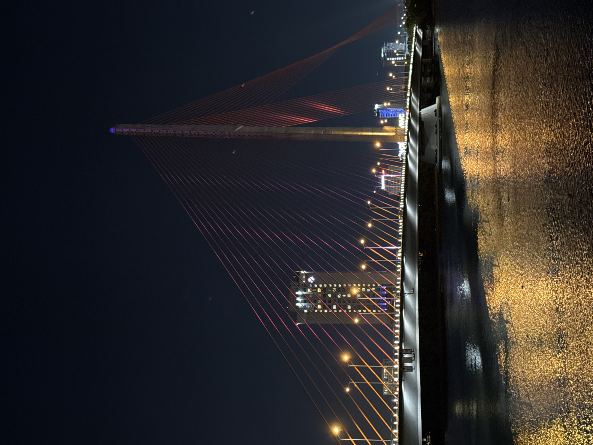This striking image captures a cable-stayed bridge illuminated at night in Đà Nẵng, Vietnam, viewed from a slightly elevated perspective. The prominent main pylon of the bridge stands on the left, topped with a distinctive purple light and adorned with white lights along its structure. Numerous slender cables, glowing in a vibrant red or pink, radiate from the pylon to connect with and support the bridge deck.
The bridge deck itself is brightly lit with a continuous array of warm yellow and white lights, creating a radiant pathway across the scene. To the right, a wide body of water, likely a river, reflects the dazzling lights of the bridge and the surrounding urban environment, forming shimmering patterns of golden and dark ripples on its surface.
In the background, behind the intricate network of cables, several modern buildings are visible. One building, partly framed by the bridge's red cables, features a grid of illuminated windows and a blue-lit sign on its facade, where the letters "WVC" are distinctly legible. The sky above is a deep, clear dark blue, providing a stark and dramatic contrast to the brilliant artificial illumination. The scene is devoid of specific human activity, emphasizing the architectural grandeur and nighttime beauty of the bridge and its illuminated surroundings in this modern city.