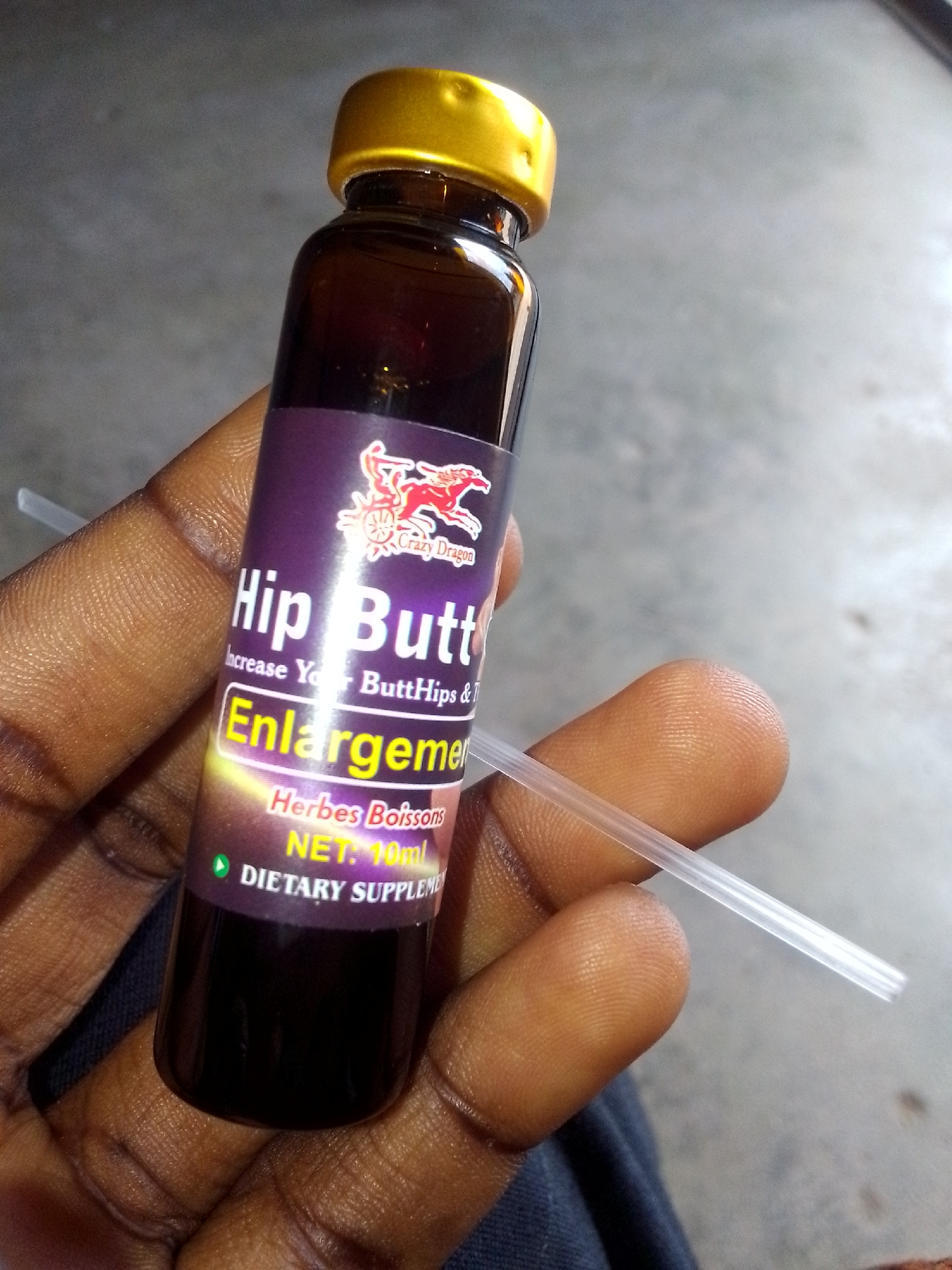 A person is holding a brown bottle with a gold cap labeled "Hip Butt Enlargement" in their hand. The bottle has a purple label with a cartoon dragon logo and text indicating it is a "Dietary Supplement." A clear, narrow plastic straw is visible next to the bottle. The person's hand and fingers are dark-skinned. The background appears to be a plain, gray surface, possibly concrete or a floor.