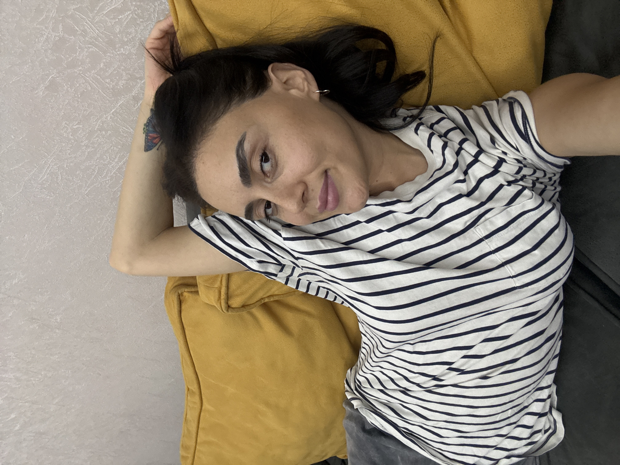 A young woman with dark hair and a butterfly tattoo on her arm is lying on a yellow sofa, looking up at the camera. She is wearing a black and white striped t-shirt and gray pants. Her head is tilted to the side, and she has a slight smile on her face. The background appears to be a textured wall. The lighting suggests it might be daytime. The scene is casual and relaxed.